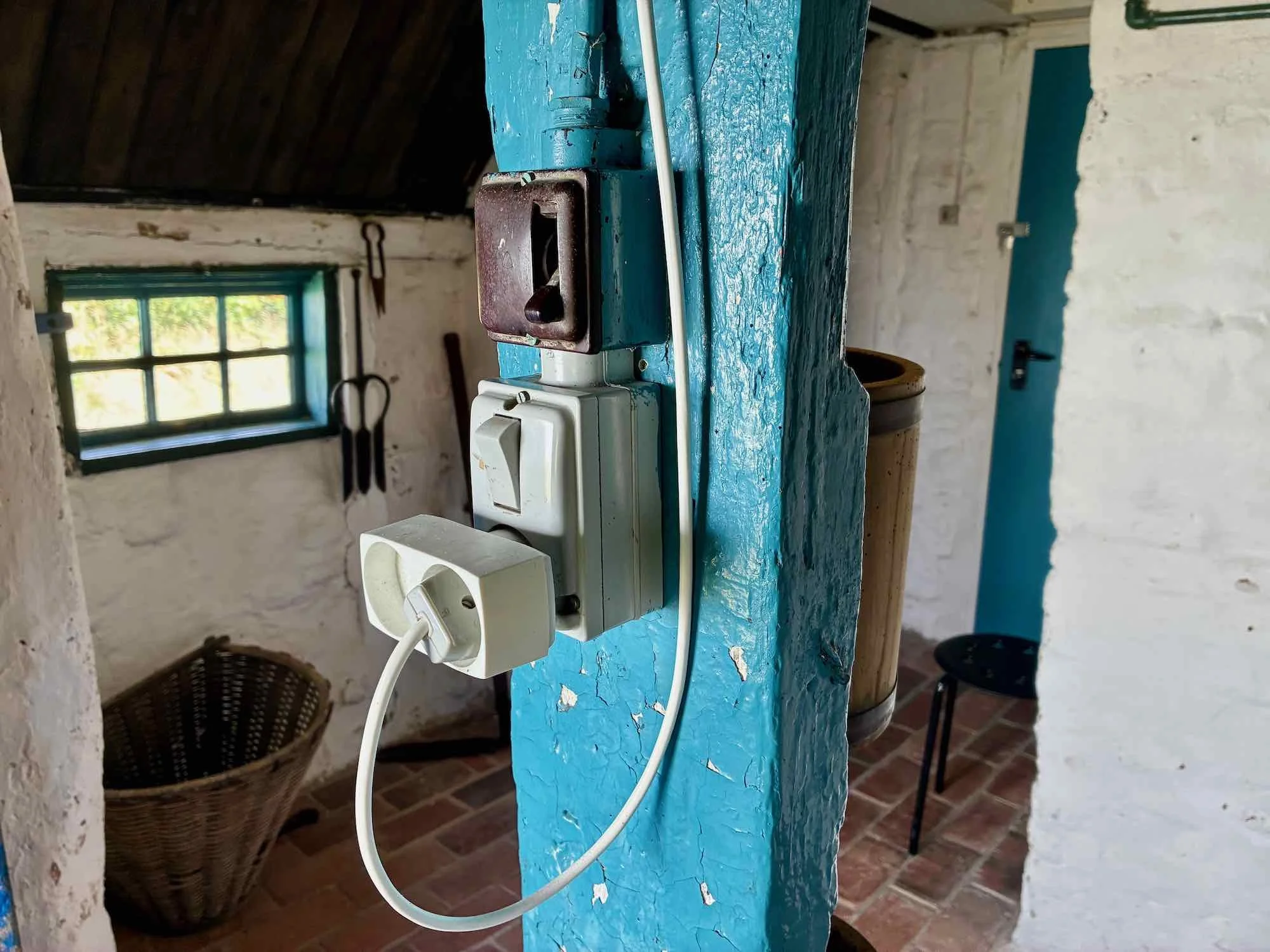 Billedet viser en elektrisk stikkontakt med ledning, der er monteret på en blåmalet væg i et gammelt rum med hvide murstensvægge og et lille vindue i baggrunden.
