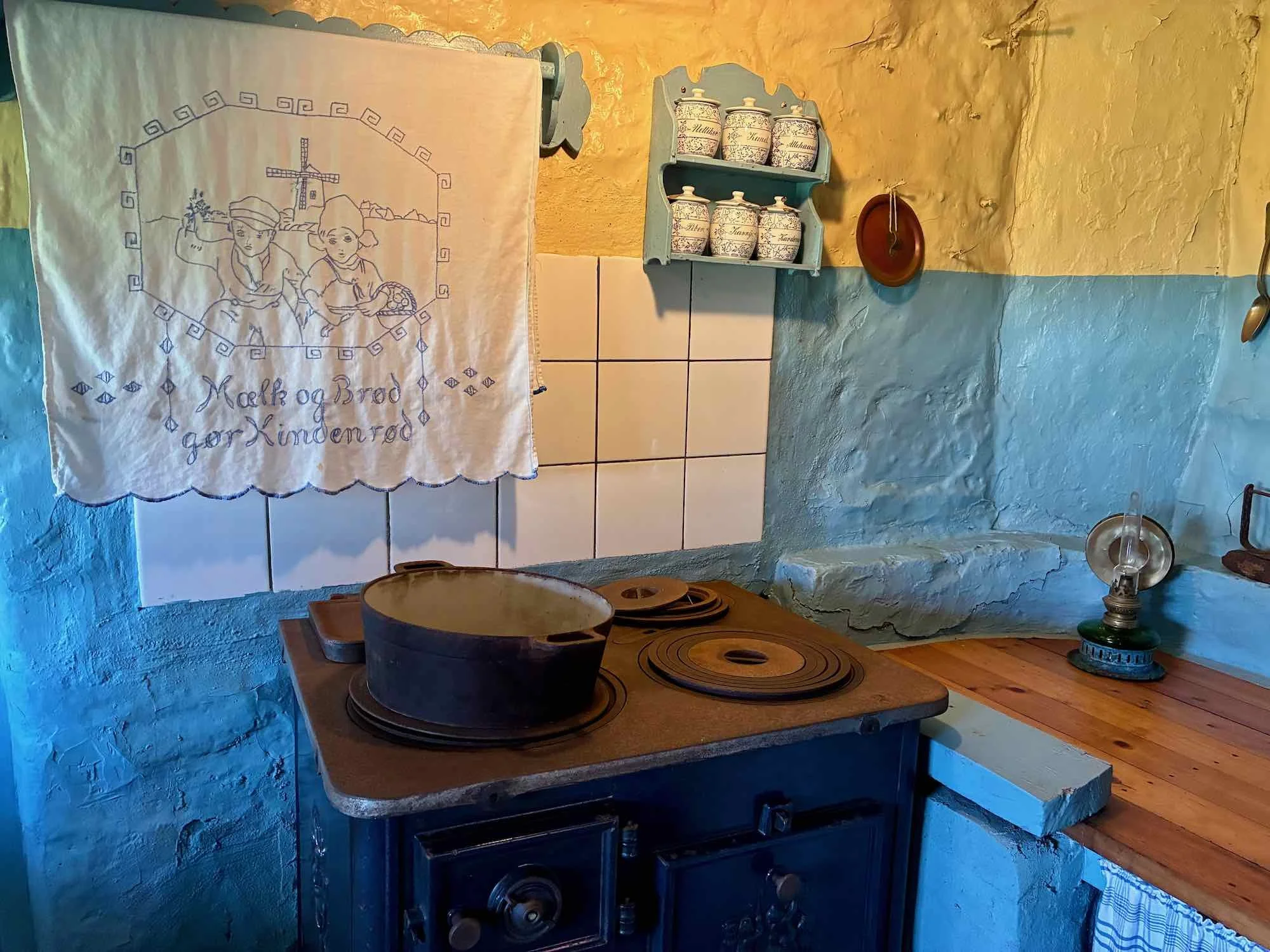 Køkken med gamle elementer, en træbordplade, en komfur med støbejernsgryde, en hvid håndklæde med blå broderi, og en hylde med syv krukker