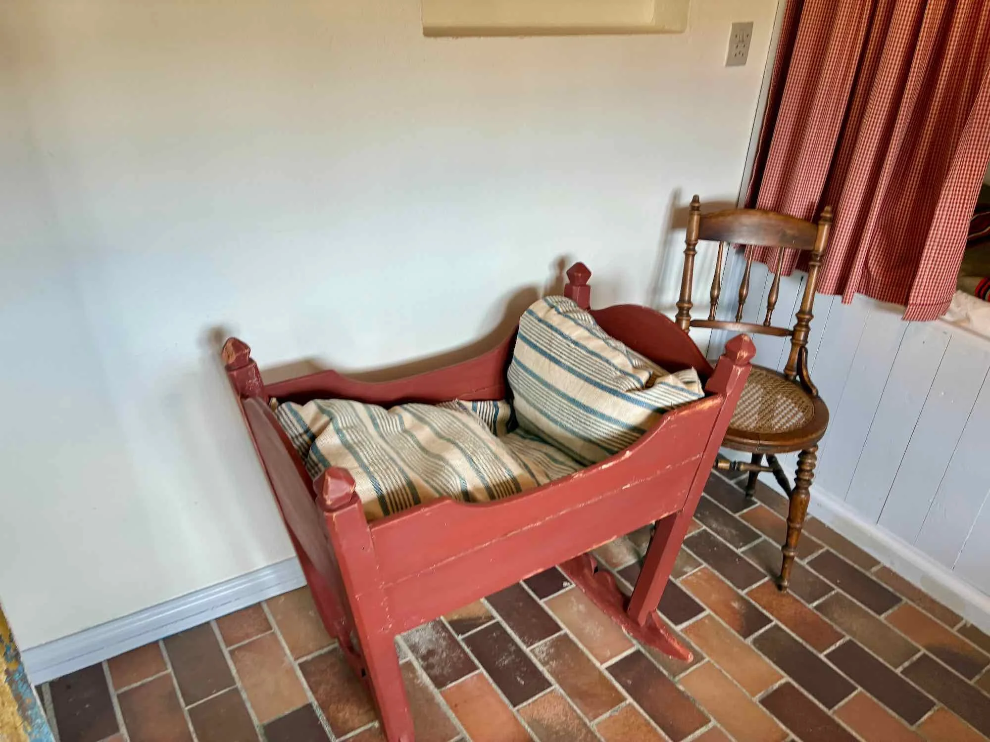 En gammel, rød tremmeseng i træ med en baby, der ligger i den, og en gammel træstol, der står bagved, ved siden af et vindue med rødt og hvidtternet gardin.