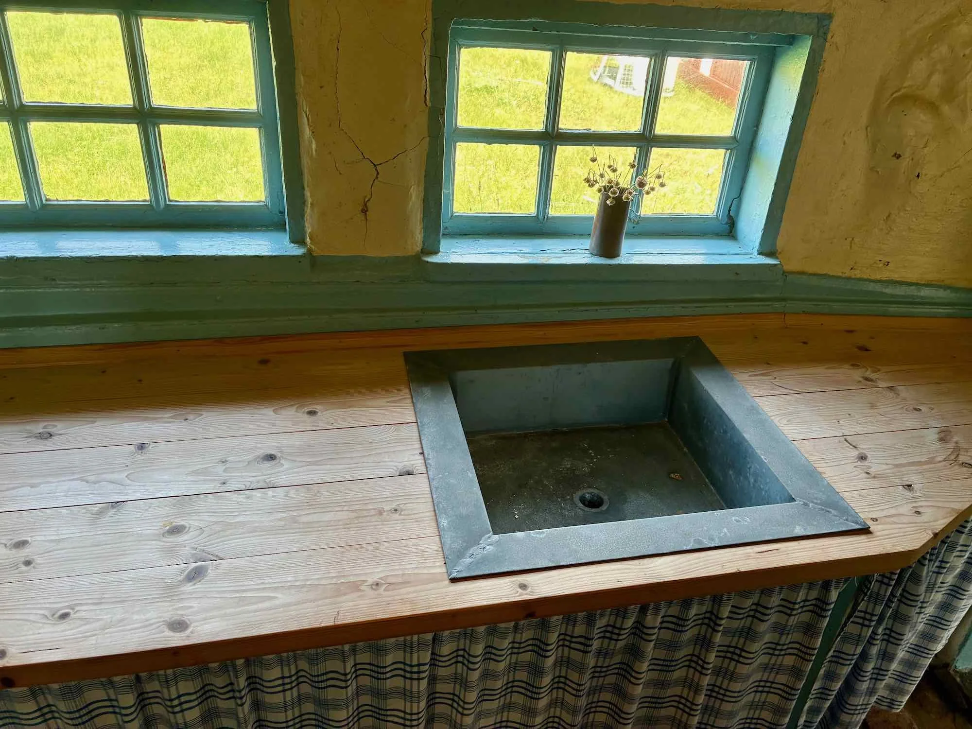 Indendørs udsigt af en lille vask eller håndvask med træbordplade foran to grønmalede vinduer med blomster i en lille vase.
