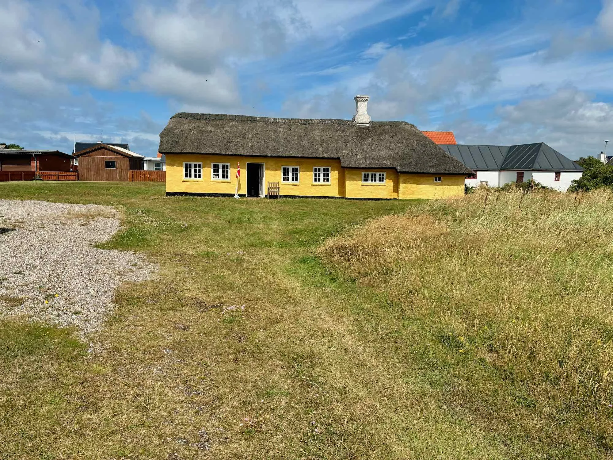 Det gamle fiskerhus i Agger med stråtag og hvid ramme rundt vinduer, omgivet af græsarealer og en grussti, under en skyet himmel.