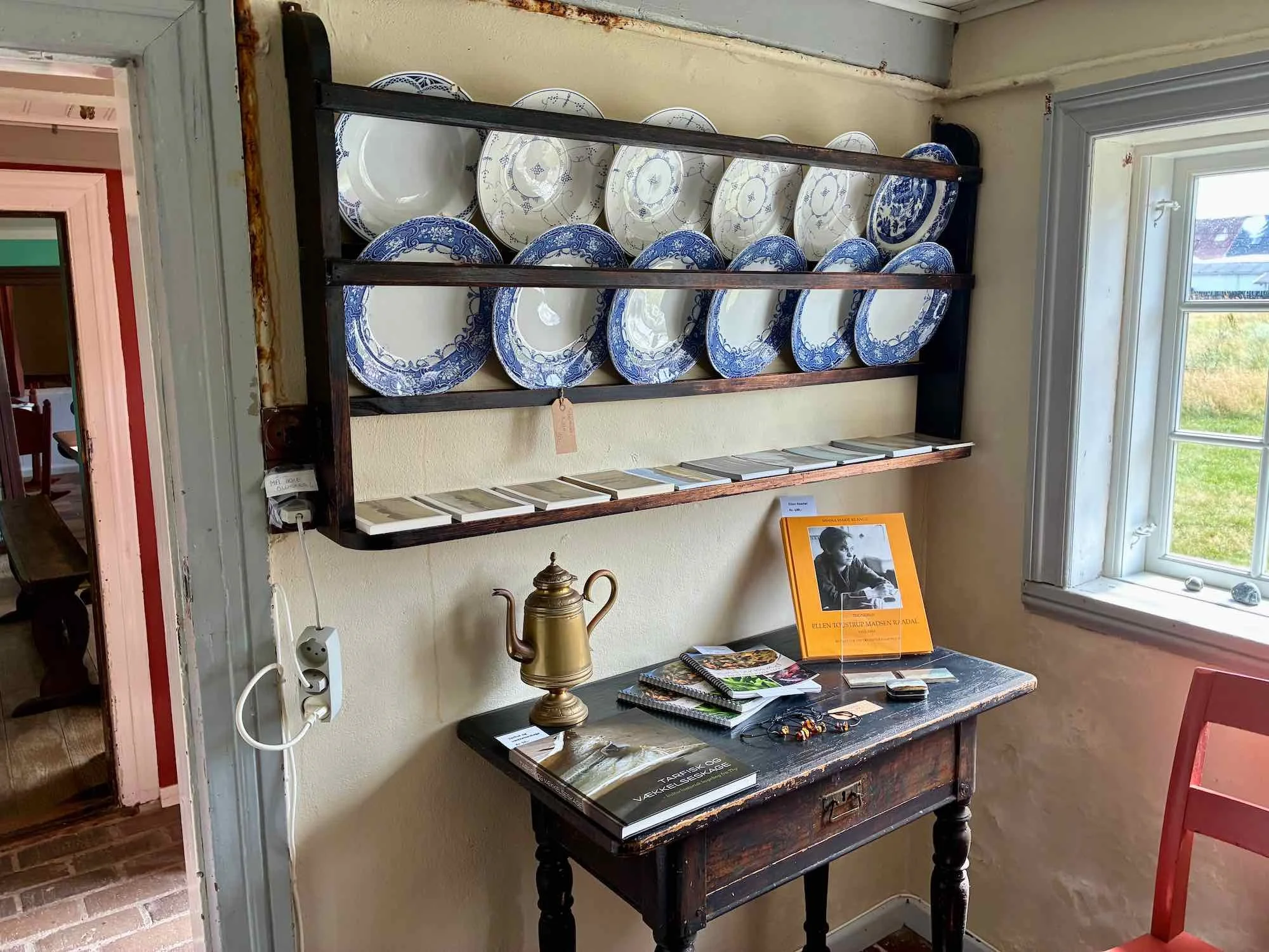 Et lille træbord under et vindue med bøger, en sølvfarvet kedel, og nogle småting. På væggen over bordet er der en sort reol med tallerkener i blå og hvid. Nær vinduet er der en stol i rød.