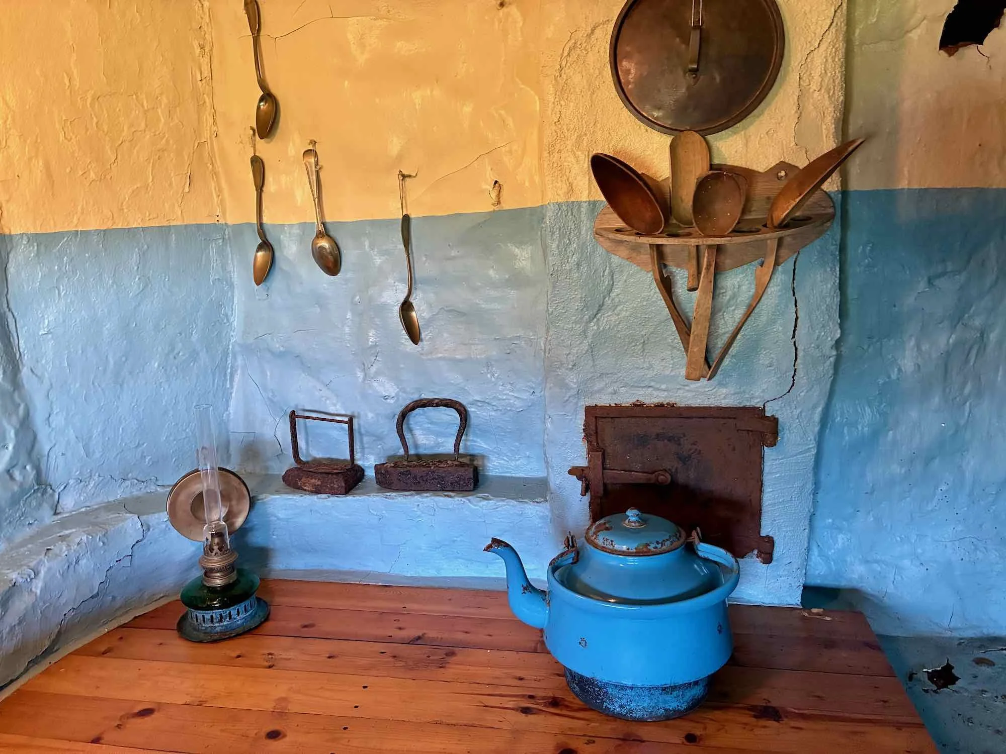 Gamle køkkenske produkter inklusive en blå kedel, jernstøtter, gamle skeer og en olielampe på en træbord.