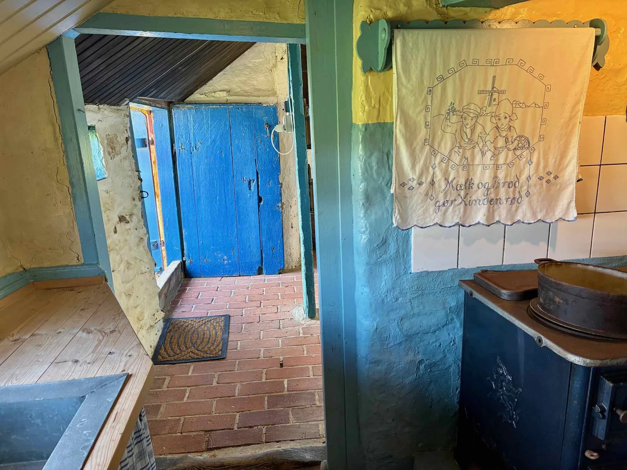 Entré med blå dør til et røgfyldt rum med kvadratisk terrakottagulv. En tekst med illustration af to kvinder og et kors, der siger 'Mekt og Brød gør Kinderneød'.