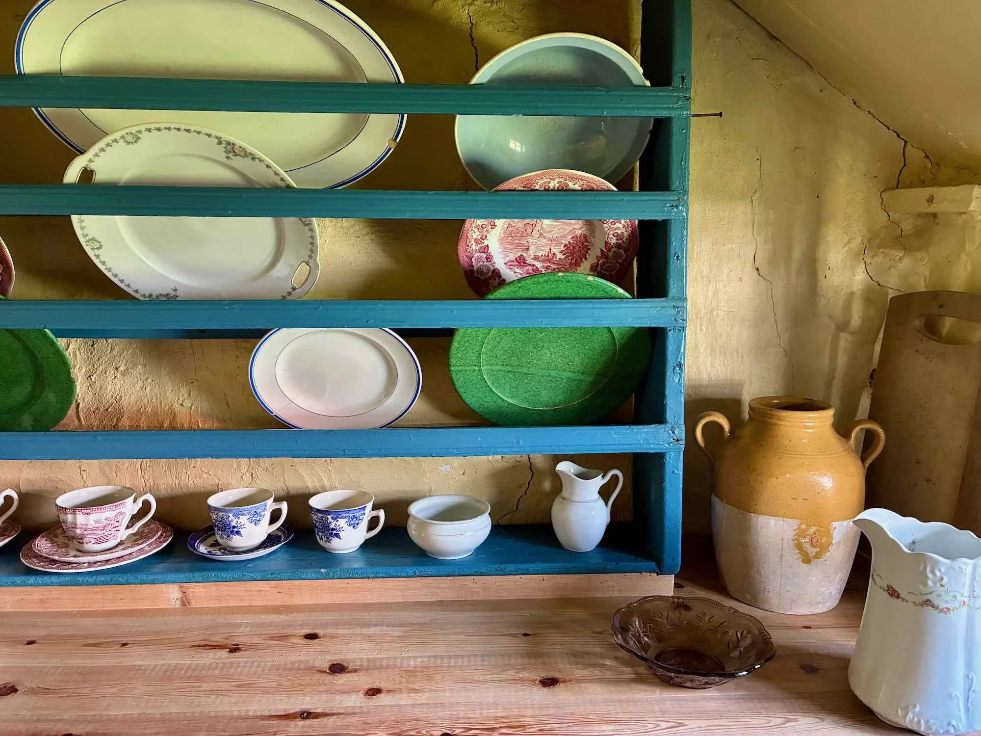 En hylde med forskellige tallerkener, kopper, karafler og krus i forskellige farver og mønstre, med en keramisk vase og en kander på bordet.