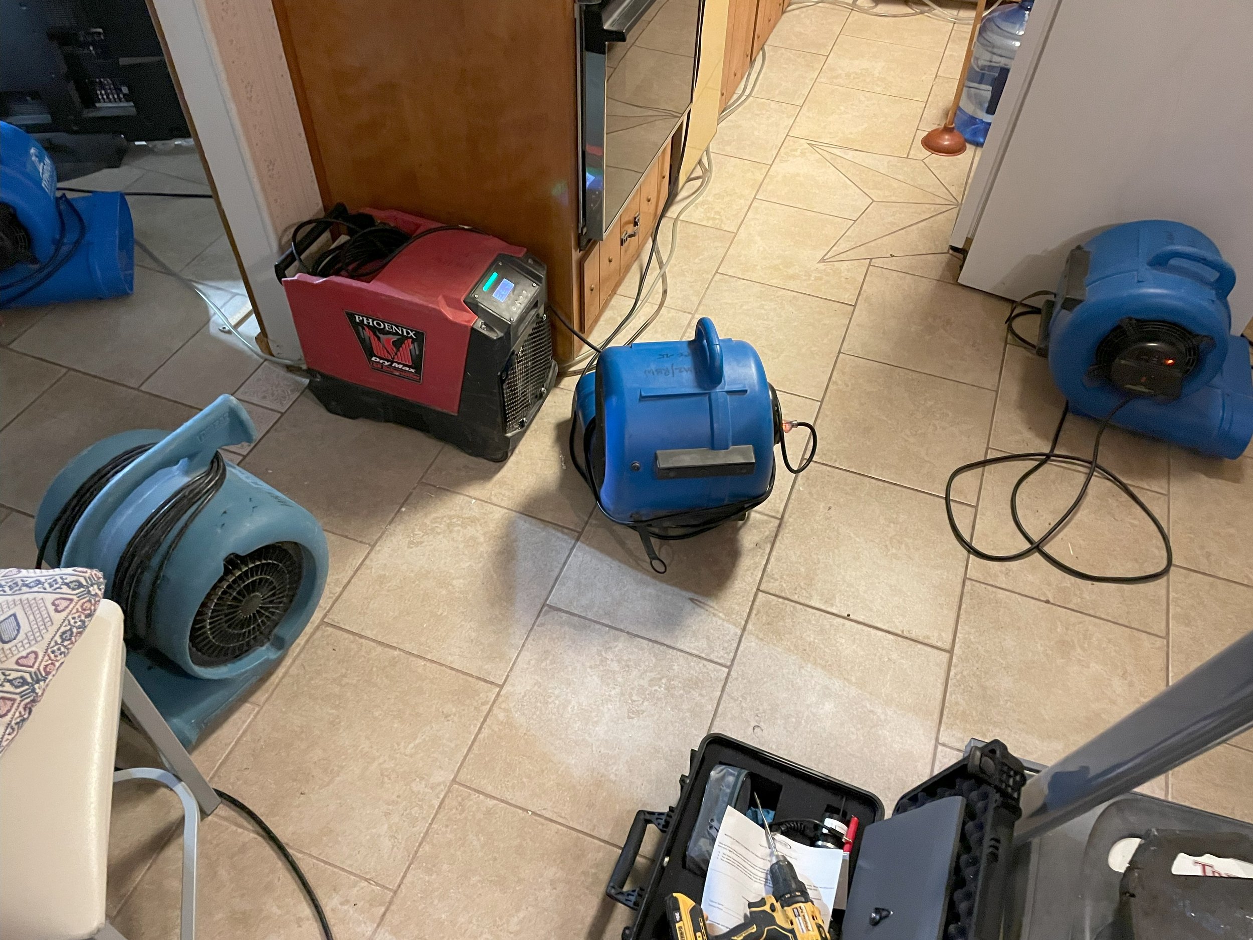 Several blue and red portable electric space heaters are placed on a tiled floor in a room. There is a ping pong type table, and various tools and equipment are nearby, including a toolbox with tools, a drill, and a large plastic water bottle.