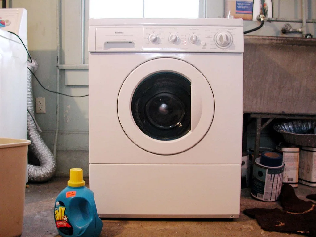 White washing machine with control knobs on top, in a laundry area with cleaning supplies and storage items around.