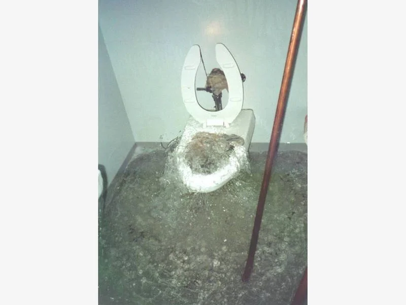 A dirty, broken toilet with the seat lid partially open, surrounded by debris and mud on the floor, with a mop leaning against the wall nearby.