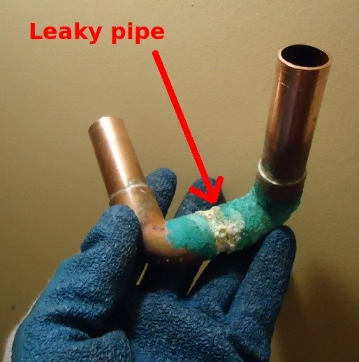 A gloved hand holding a metallic pipe with a leaky joint, marked with a red arrow and label reading 'Leaky pipe.'