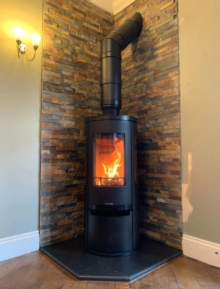Black freestanding wood stove with glass door showing flames, set on a black hearth, against a wall with stacked stone decor, with a sconce light on the left wall.