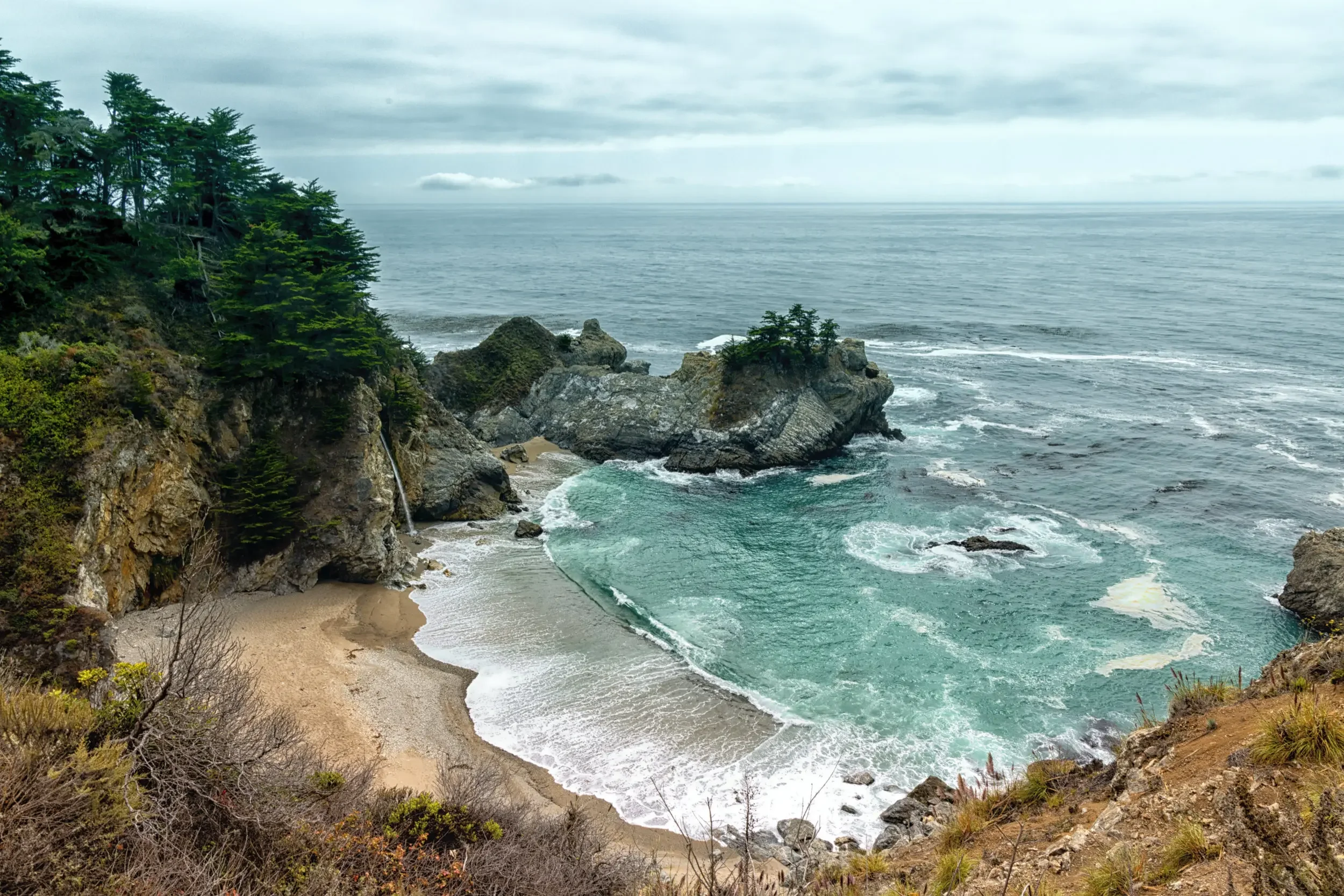 A scenic cove with a sandy beach, rocky cliffs, and evergreen trees, overlooking the ocean with overcast skies.