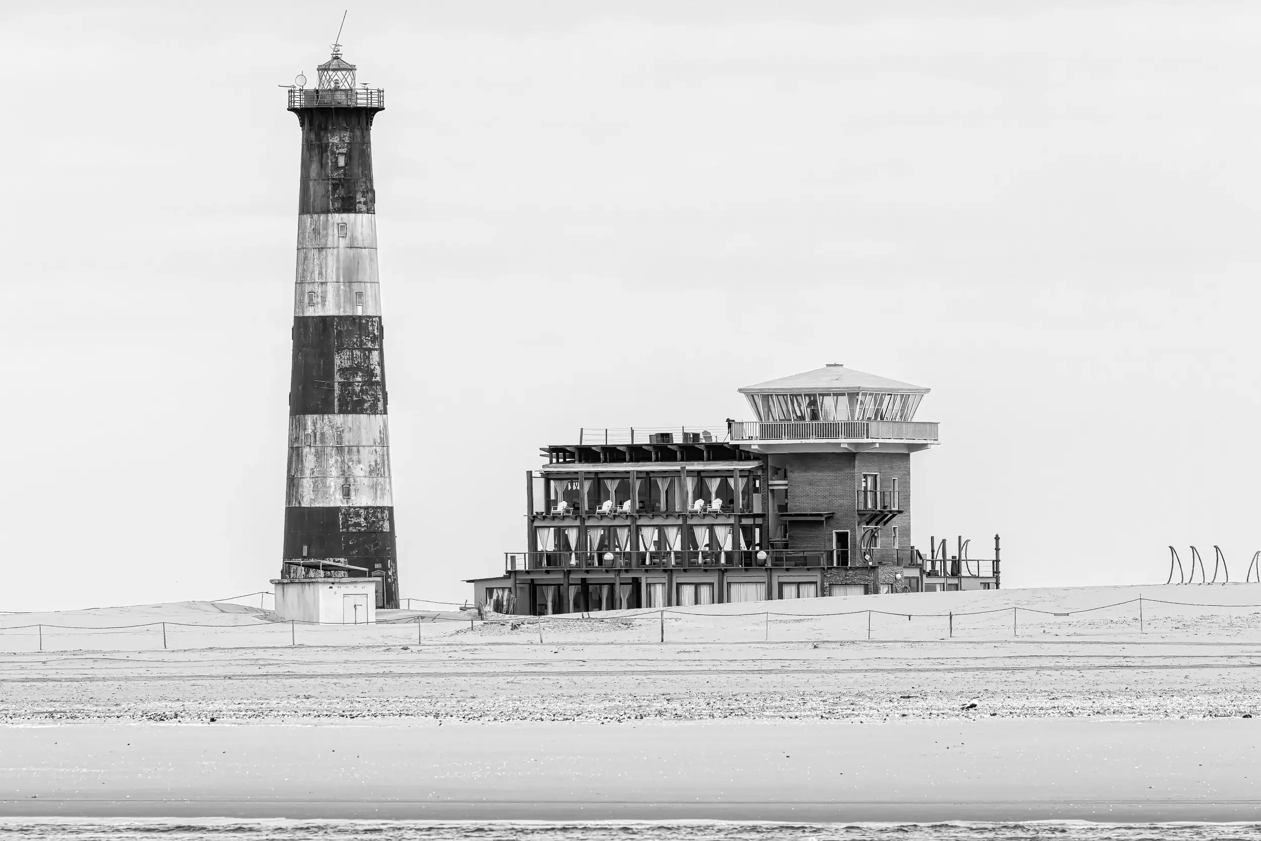 Black and white photo of a lighthouse next to a modern building on a beach.