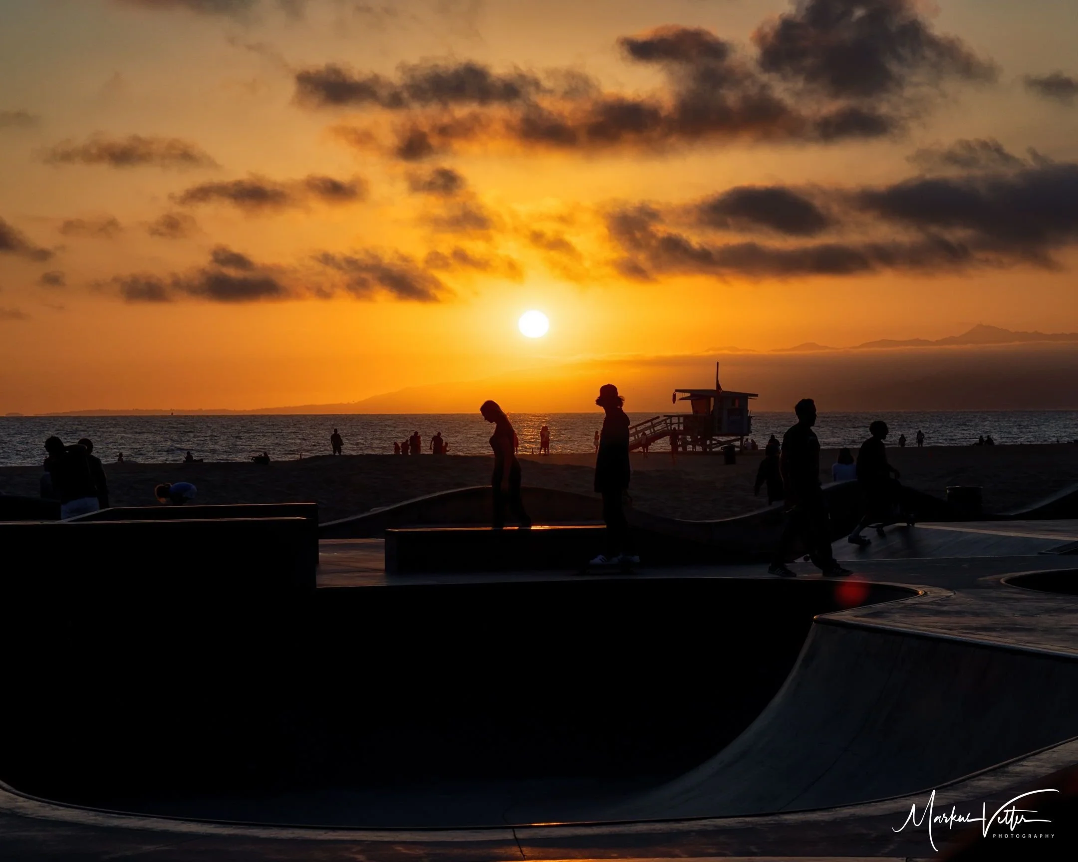Menschen, die einen Skatepark bei Sonnenuntergang am Strand besuchen, mit dunklen Wolken am Himmel und dem Meer im Hintergrund.