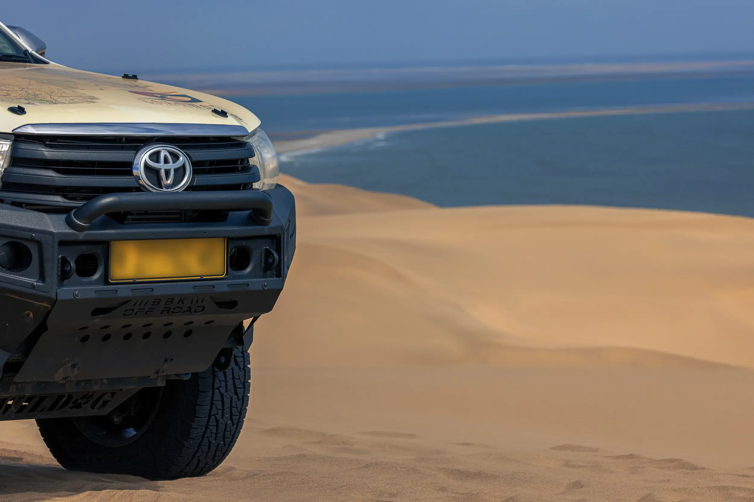 Front view of a Toyota off-road vehicle parked on sandy dunes with an ocean in the background.