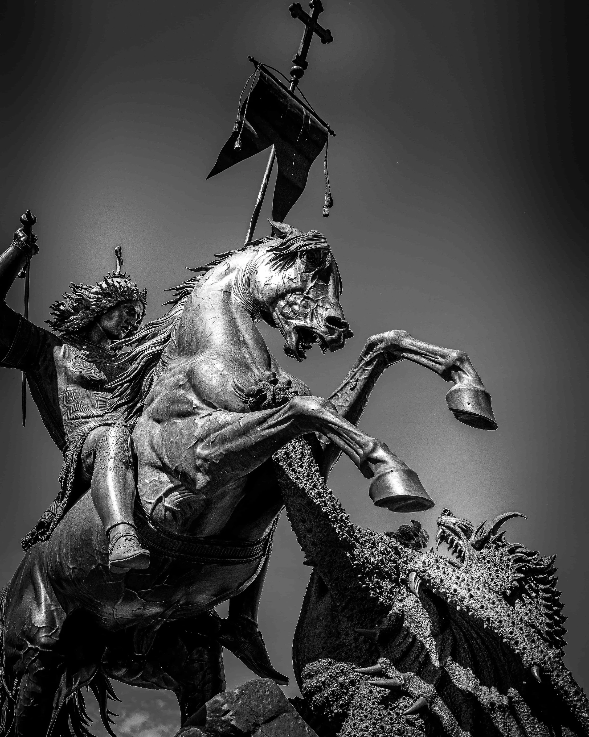 Black and white photograph of a statue depicting a warrior riding a rearing horse, wielding a sword and shield, with a dragon beneath the horse.