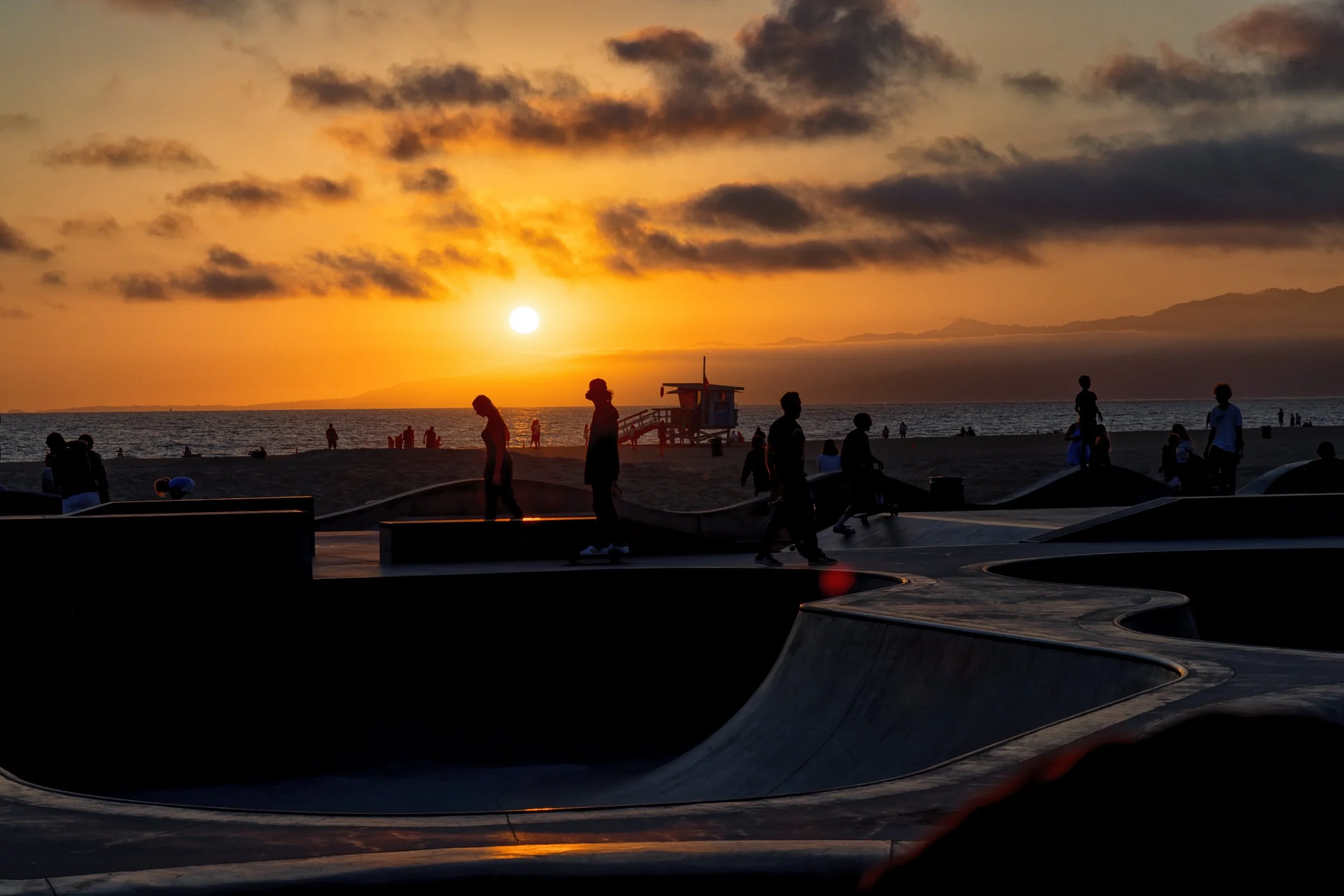 Silhouettes of people skateboarding and walking at a skatepark during sunset over the ocean.