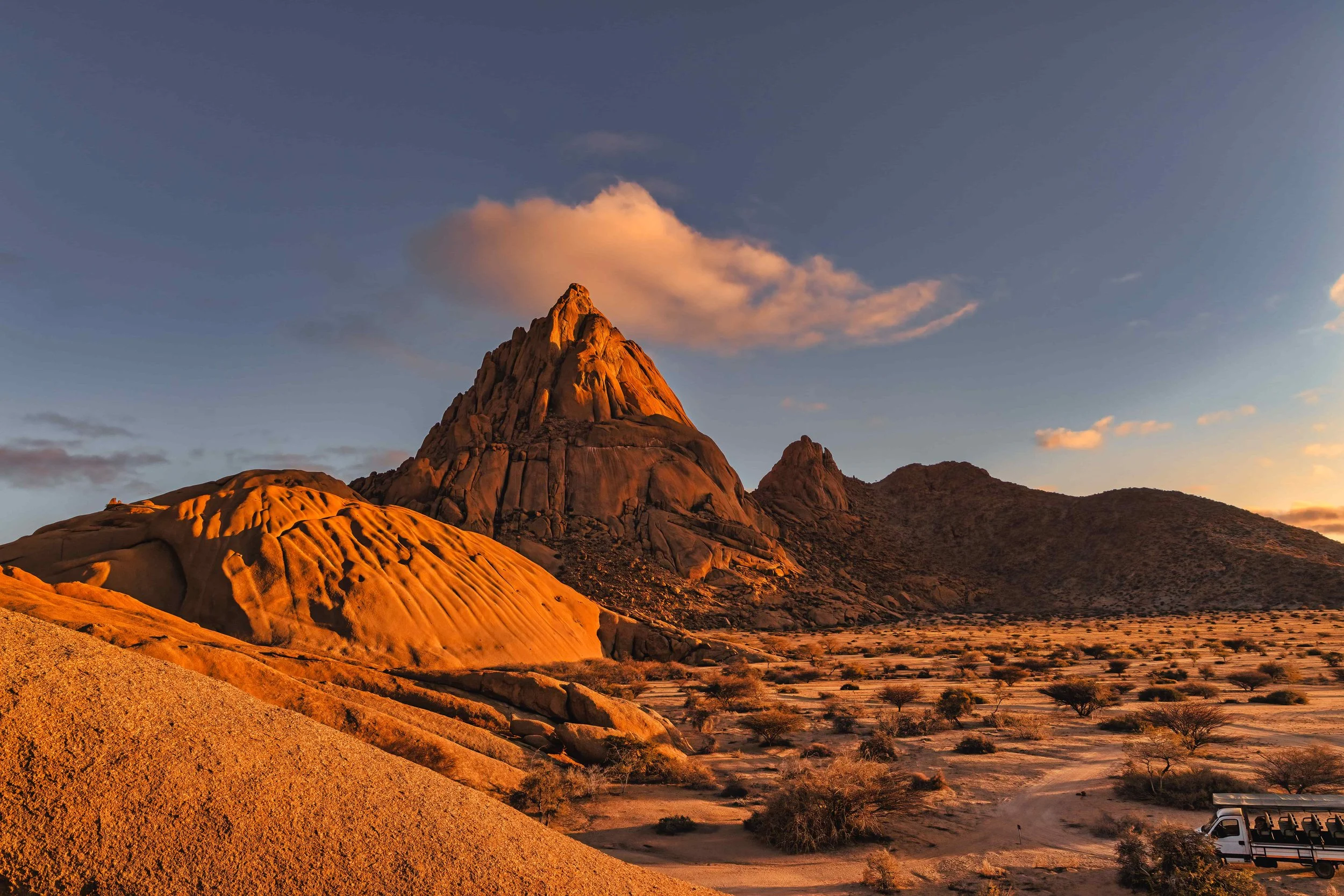Felsformationen in einer Wüstenlandschaft bei Sonnenuntergang mit vereinzelten Büschen und einem Reisebus.