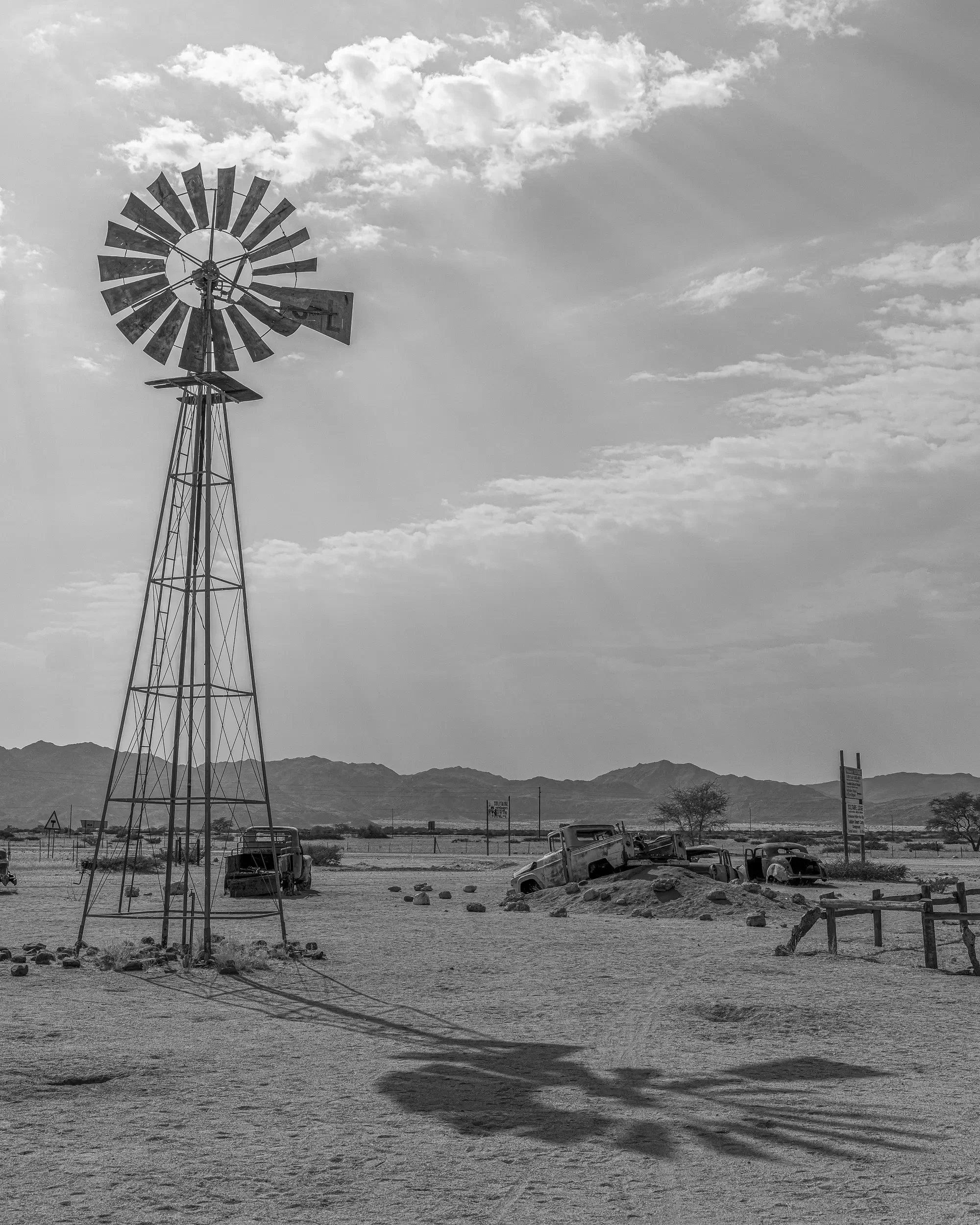 A black and white landscape featuring an old windmill, abandoned cars partially buried in sand, distant mountains, and sparse desert vegetation under a partly cloudy sky.