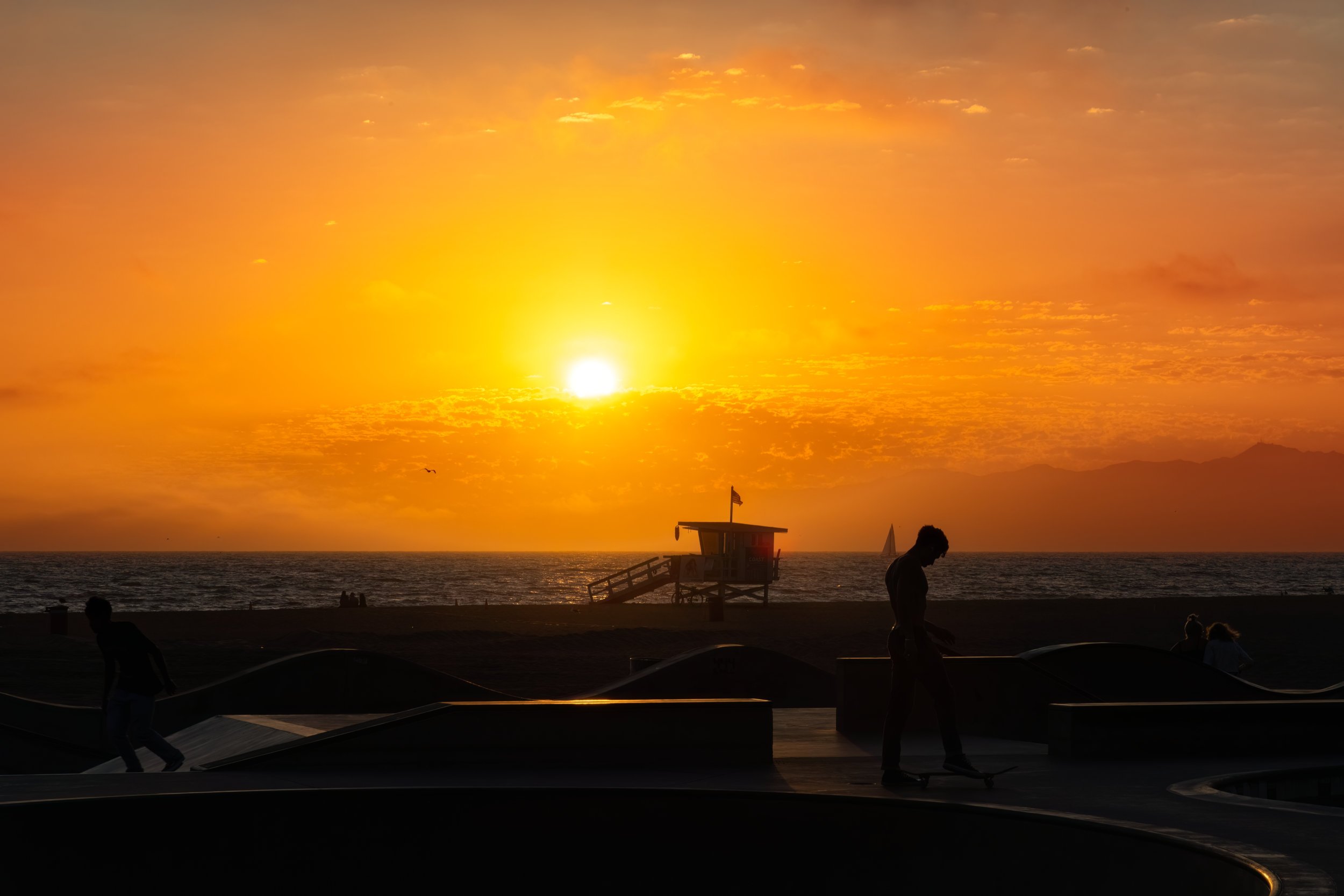 Silhouettes of people at a skate park during a sunset by the ocean, with a lifeguard tower on the beach and a sailboat on the water.