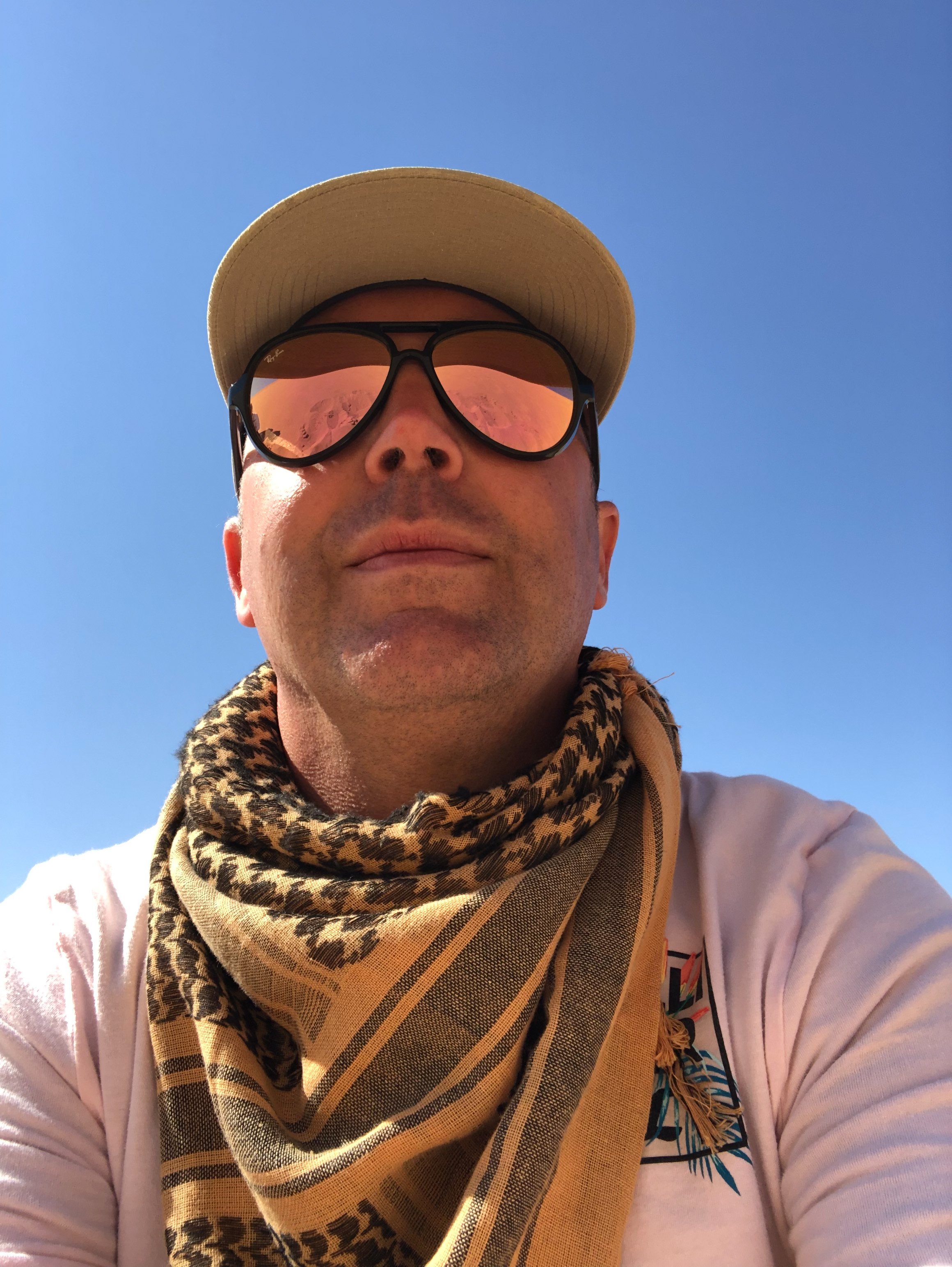 Nahaufnahme eines Mannes mit Sonnenbrille, Basecap, und Schals, der nach oben blickt, bei blauem Himmel.