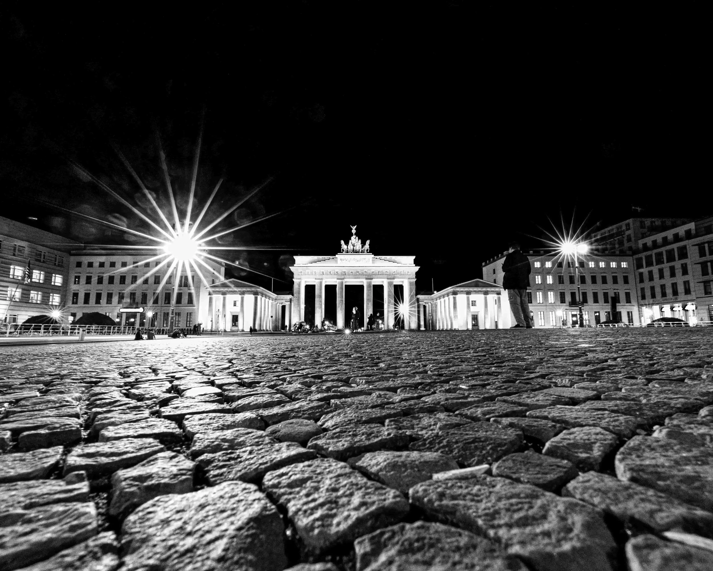 Nighttime view of the Brandenburg Gate in Berlin, illuminated and viewed from a low angle with cobblestone pavement in the foreground and bright streetlights creating starburst effects.