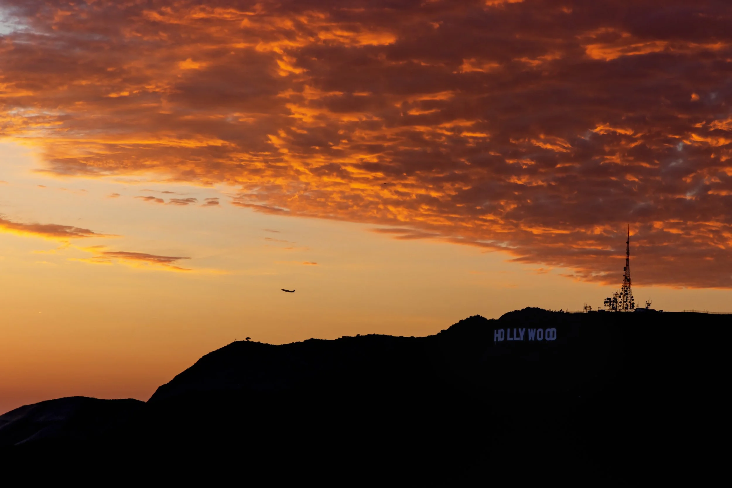 Sunset with orange clouds over Hollywood Hills, with the Hollywood sign partly visible and an airplane flying in the sky.