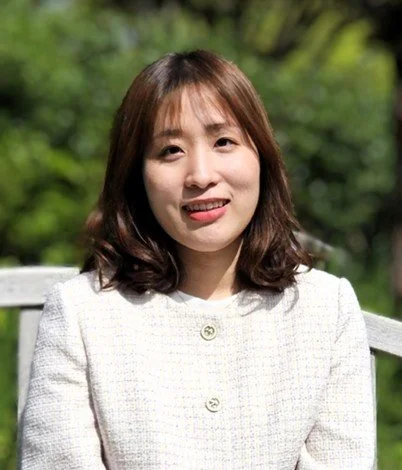 A woman sitting outdoors on a white bench with green trees in the background, smiling at the camera.