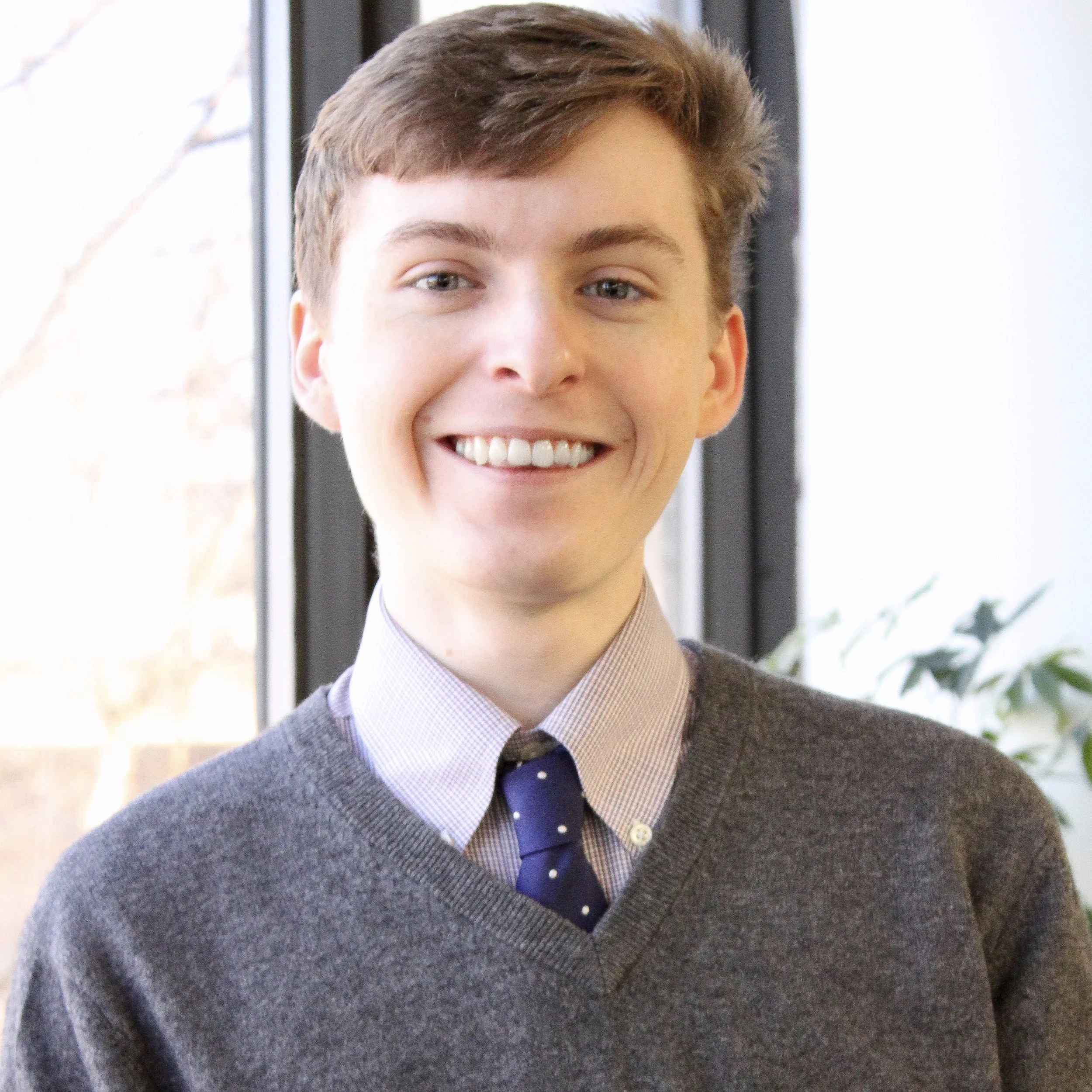 A young man with short brown hair, smiling, dressed in a gray sweater over a collared shirt with a dark blue tie with white polka dots, standing indoors near large windows.
