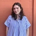 Young woman with long dark hair, wearing blue and white striped shirt, standing in front of a brick wall.