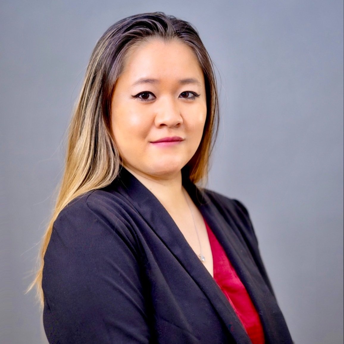 A portrait of a woman with long light brown hair, wearing a black blazer over a red top, standing against a plain gray background.
