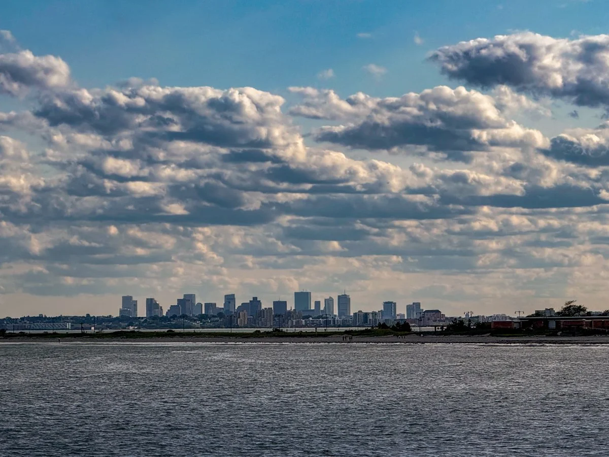 Boston skyline seen from north shore coastline with puffy clouds and blue sky overhead.