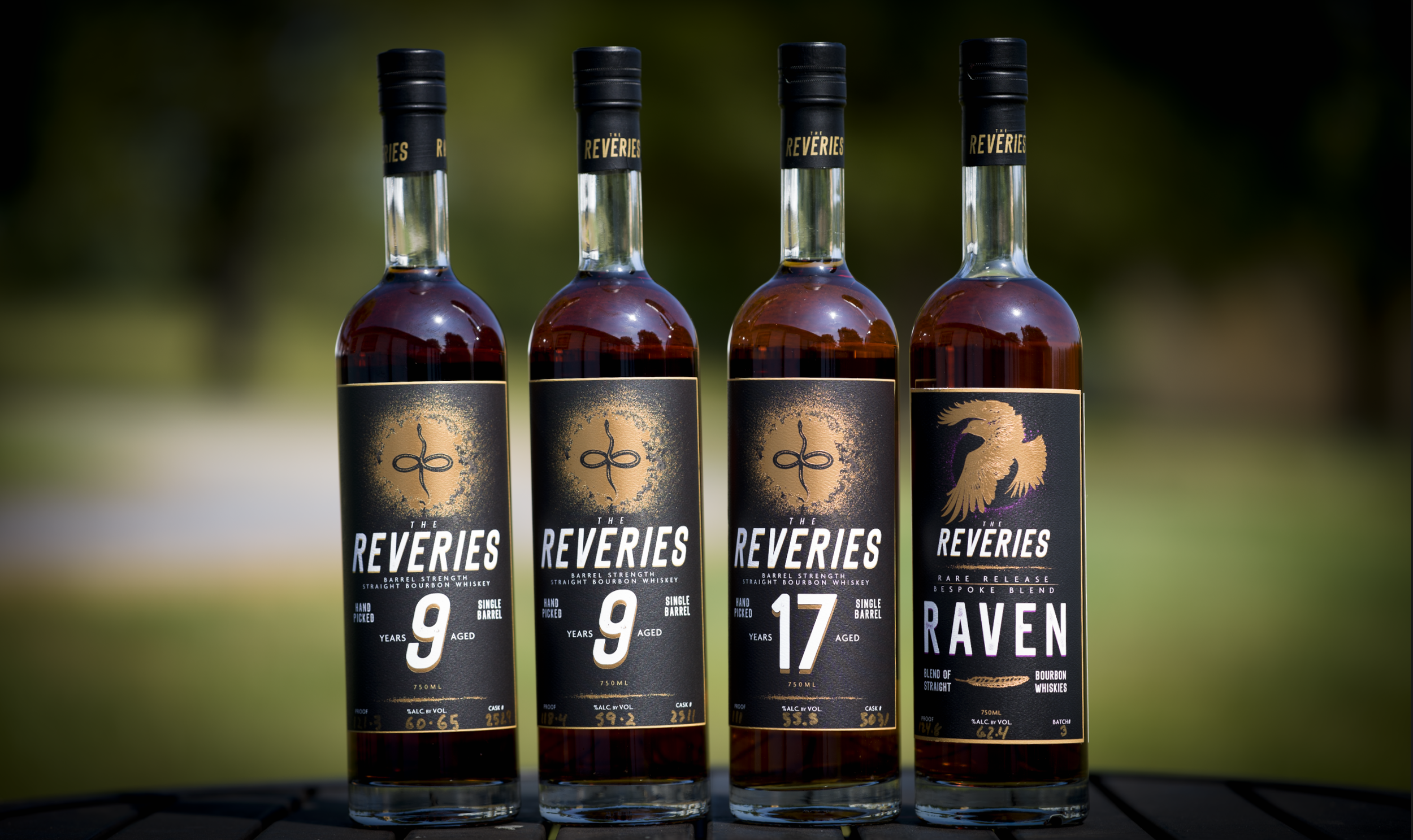 Four bottles of bourbon whiskey labeled The Reveries, with one labeled Raven, placed outdoors on a wooden surface, with a blurred green background.