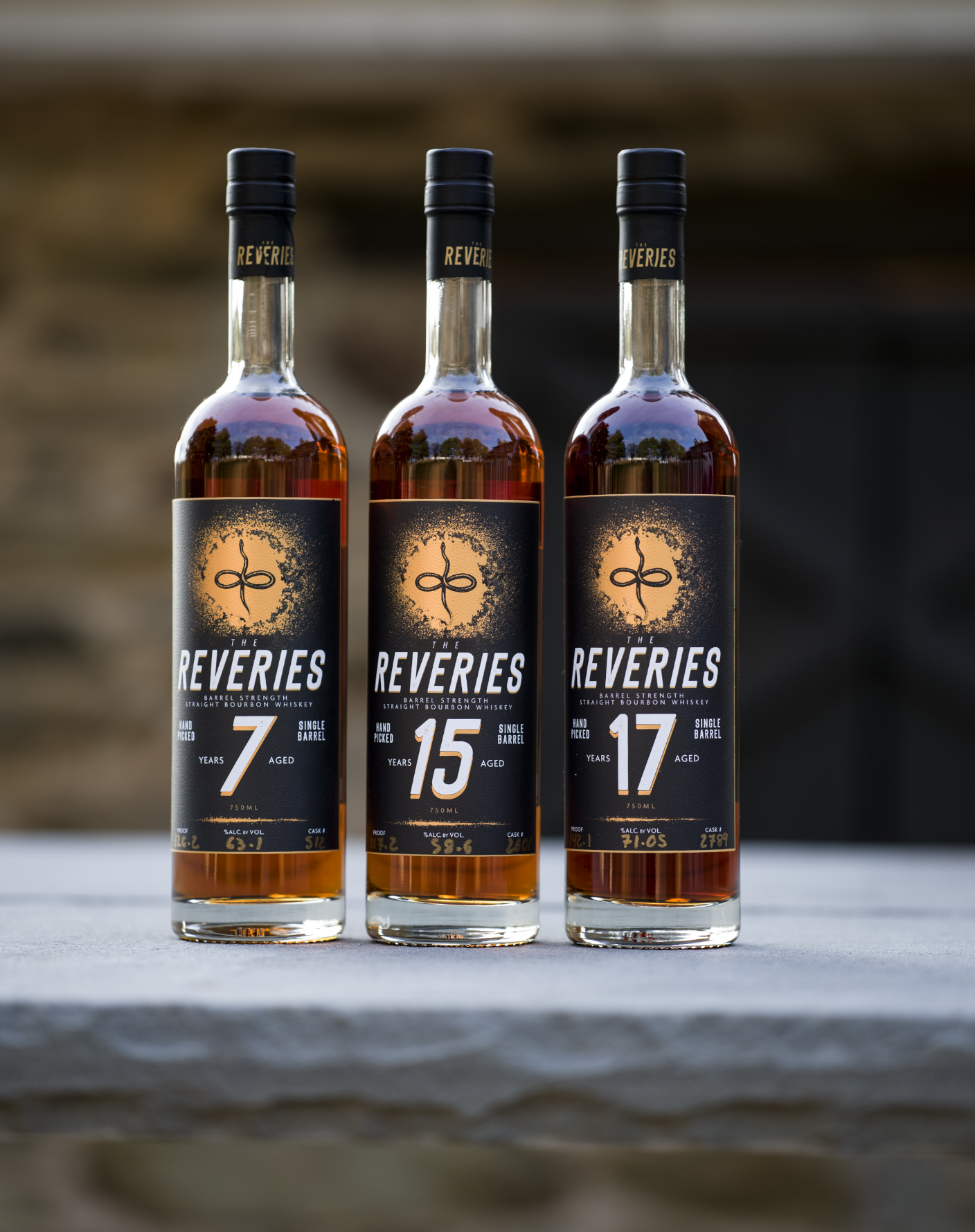 Three bottles of The Reveries bourbon whiskey, aged 7, 15, and 17 years, standing on a surface outdoors.