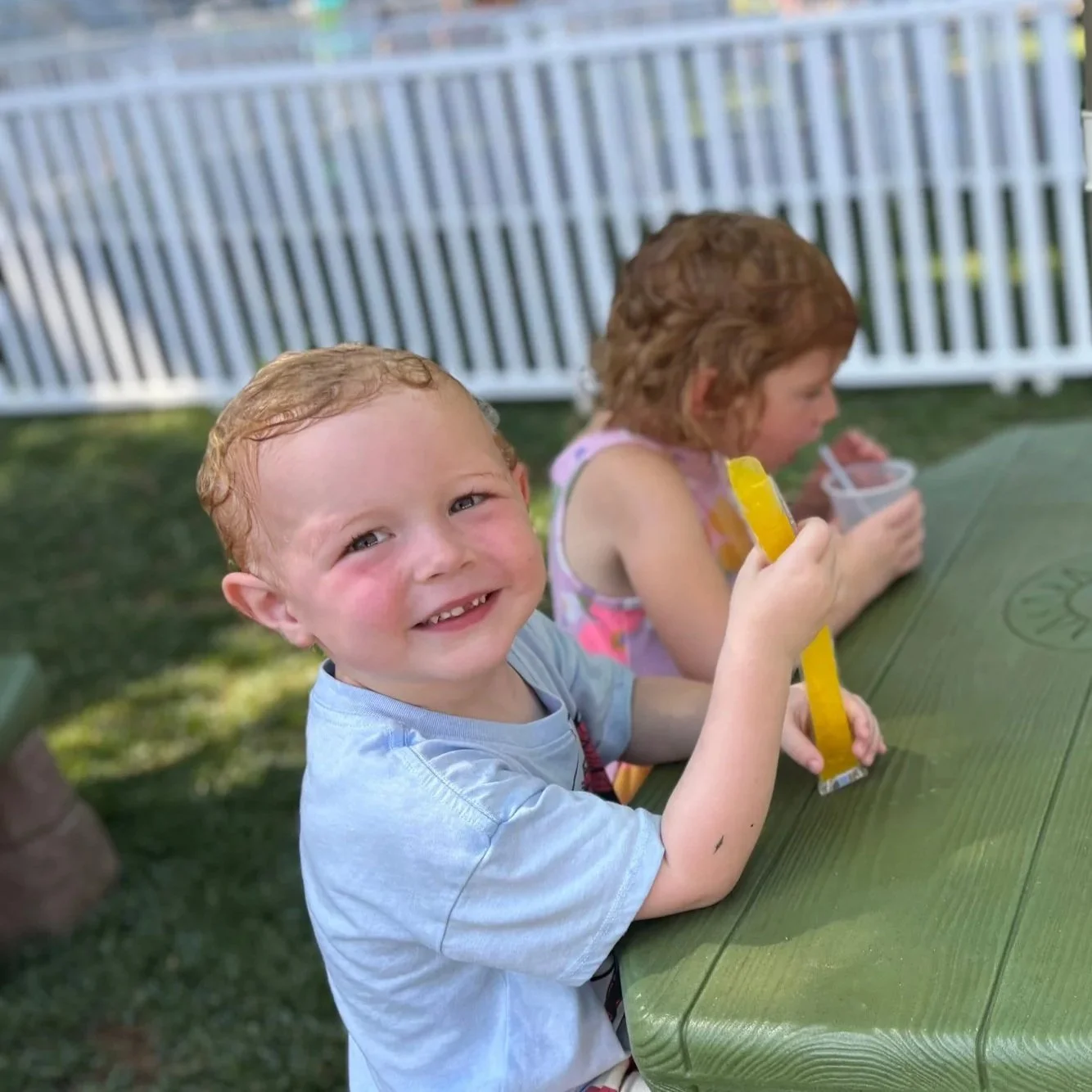 Preschool children playing outside in a sand box with various construction toys and sand toys. There is a climbing wall in the background in a wooded setting.