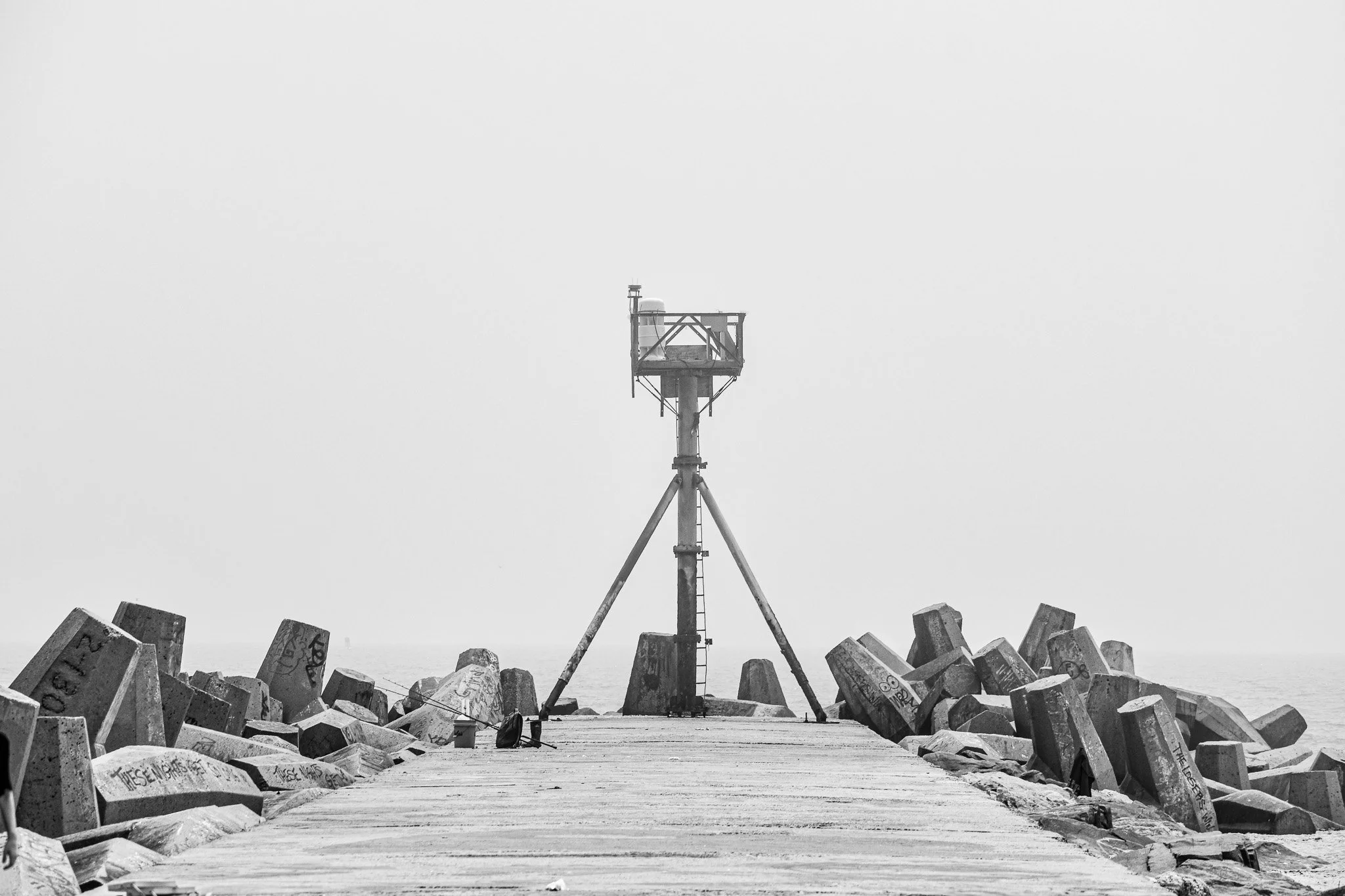 Black and white photo of a concrete pier with a metal structure at the end, surrounded by large angular rocks or breakwaters, with a calm sea in the background.