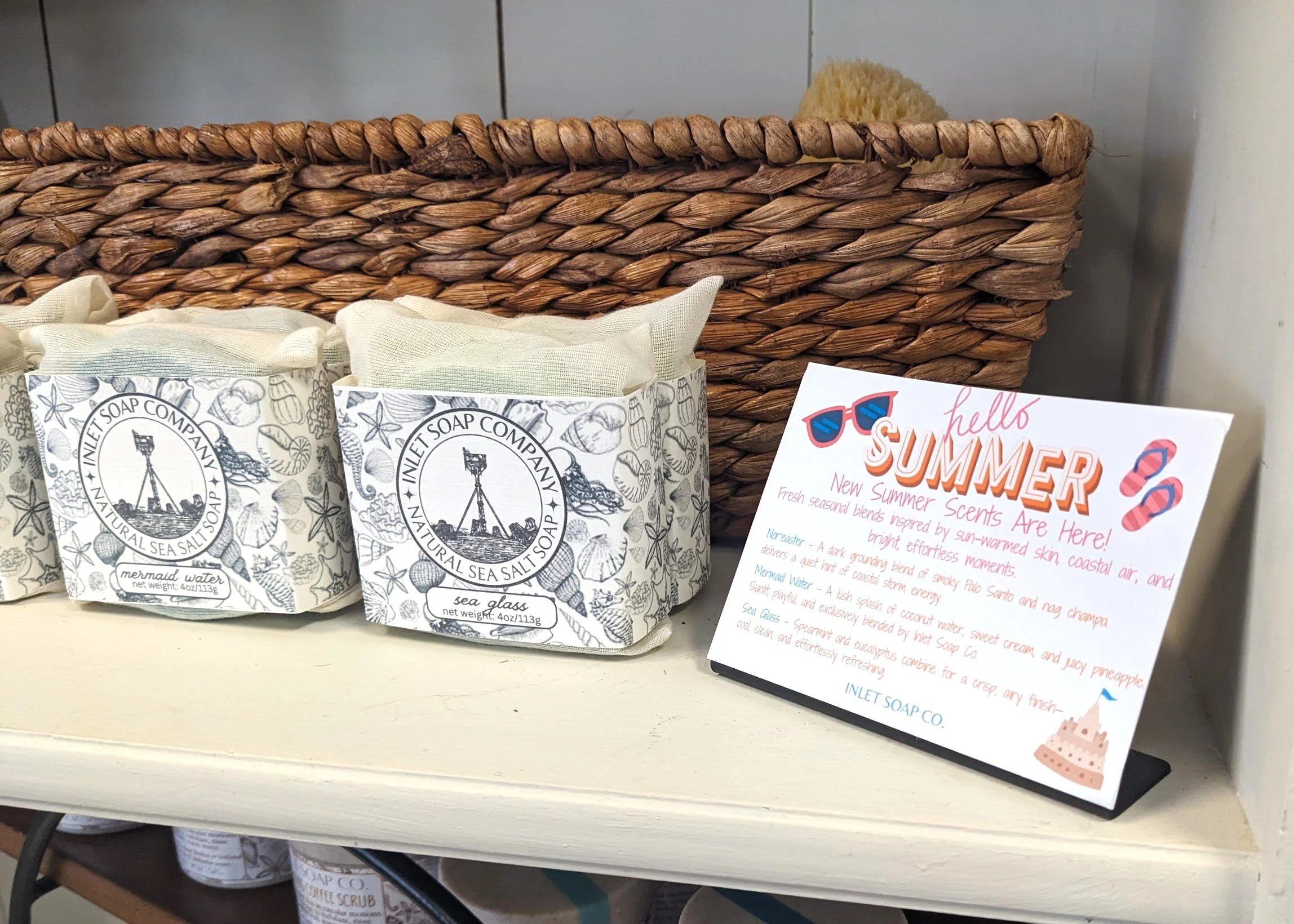 Display of sea salt soap bars with black and white labels featuring a lighthouse and seashell illustrations, and a colorful sign promoting summer scents with sunglasses, flip-flops, and a cake graphic.