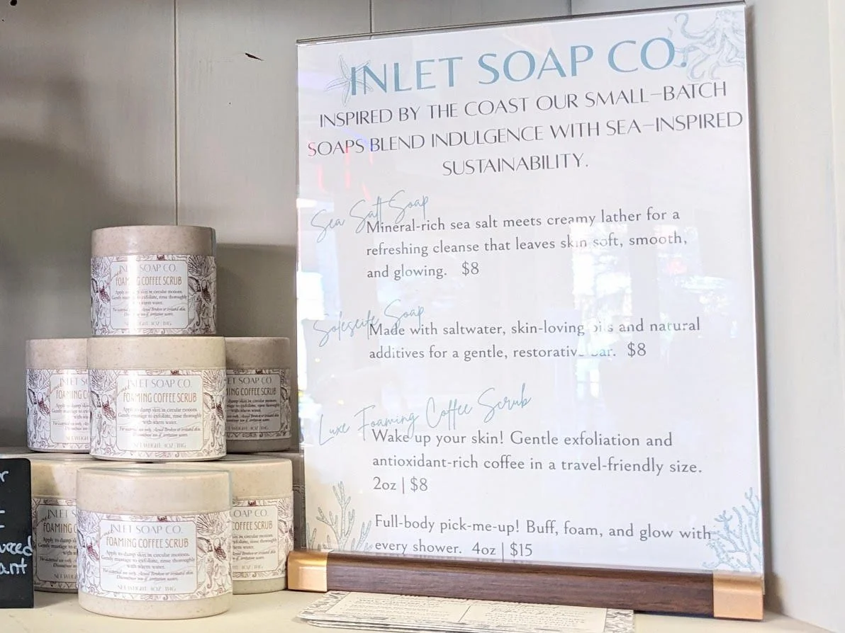 Several containers of Inlet Soap Co. foaming coffee scrub stacked on a surface next to a menu sign listing soap and scrub products with descriptions and prices.