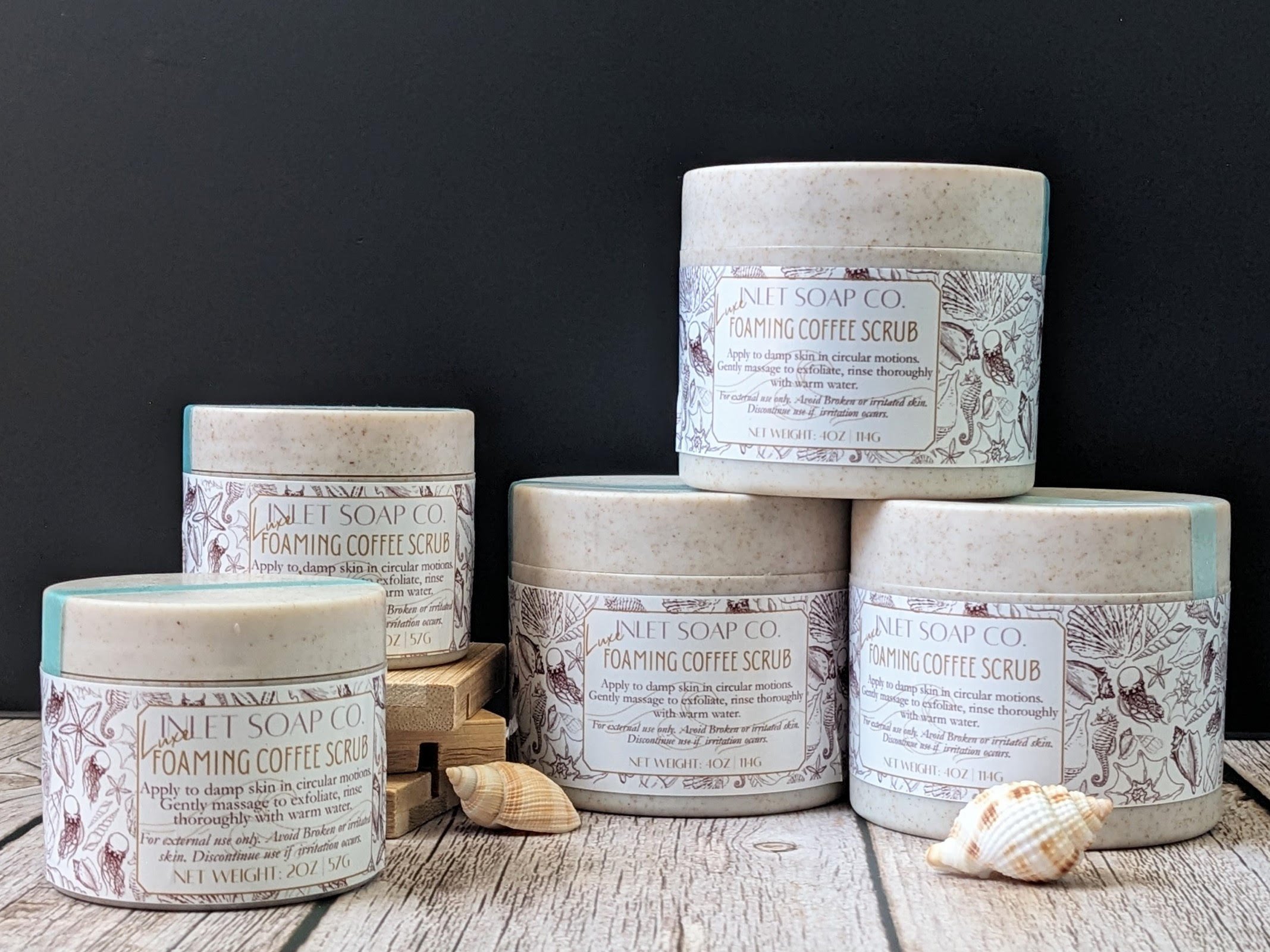 Multiple containers of foaming coffee scrub from Lulet Soap Co., arranged on a wooden surface with seashells, against a black background.