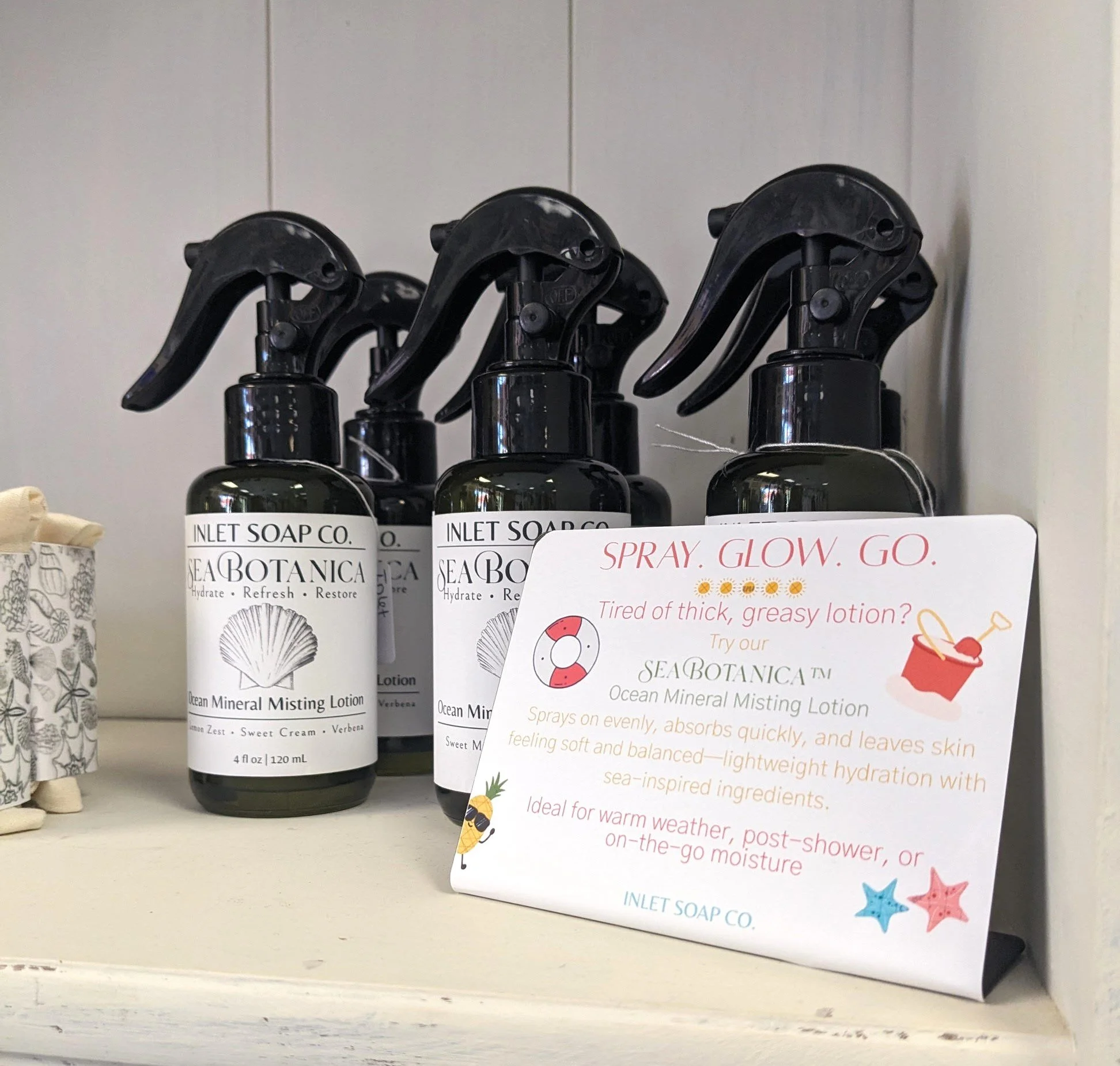 Several bottles of Sea Botanica Ocean Mineral Misting Lotion with spray nozzles, placed on a retail shelf, and a product information card with illustrations of pineapple and starfish.
