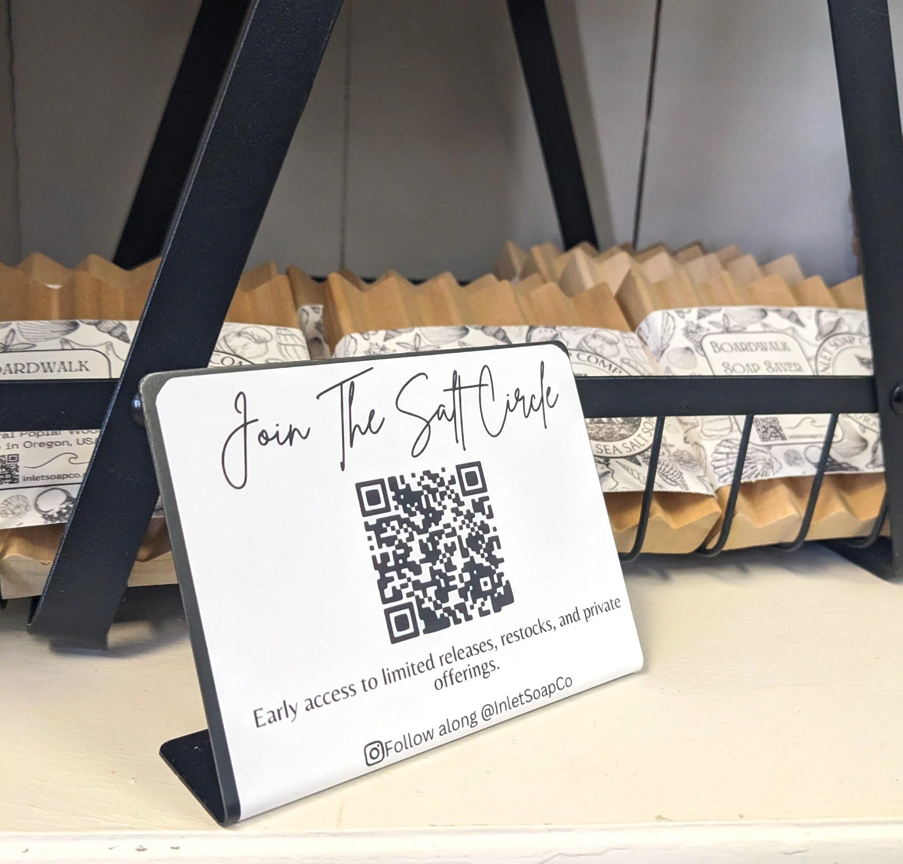 A sign encouraging people to join a salt circle with a QR code, placed on a table in front of a black metal basket filled with packaged soap products.