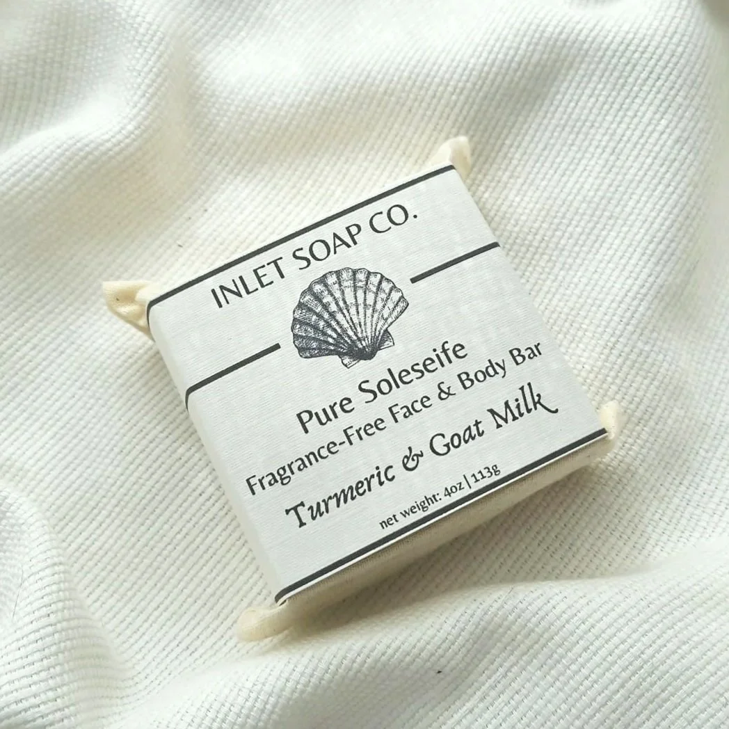 A bar of soap labeled 'Pure Soleseife' with fragrance 'Turmeric & Goat Milk' from Inlet Soap Co., wrapped in white paper with shell illustration, resting on a textured white surface.