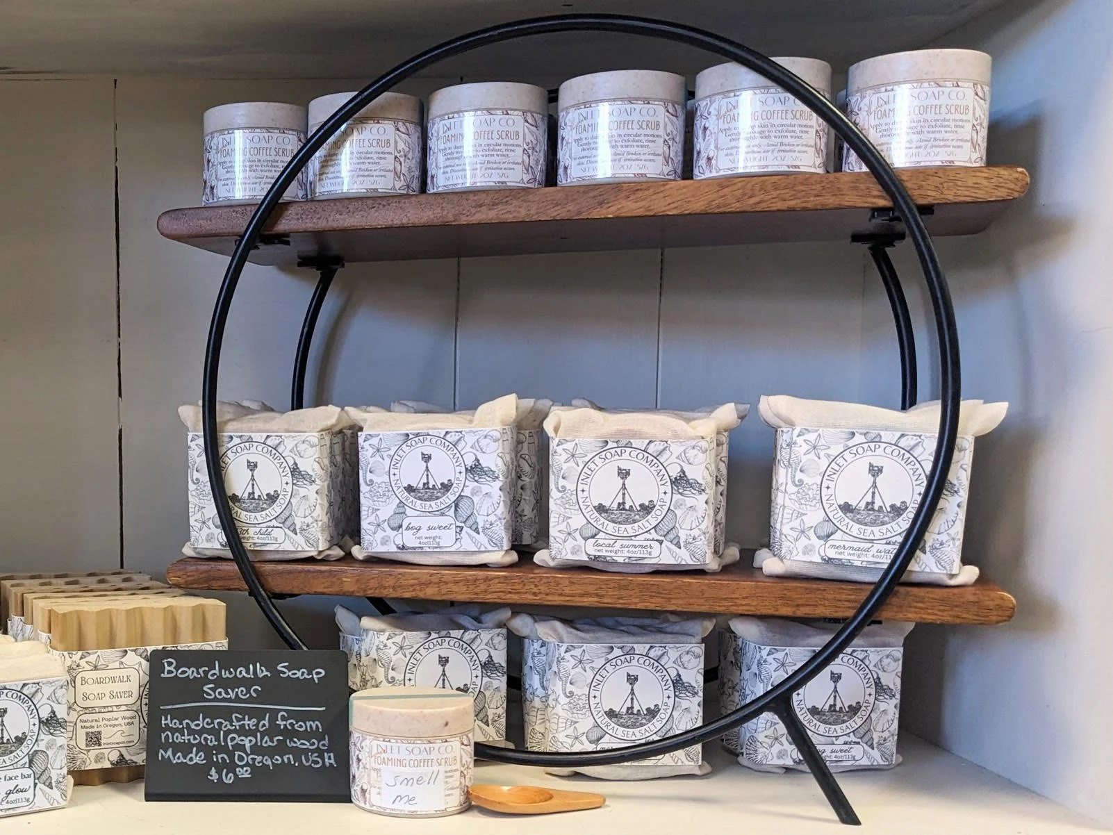 Shelf displaying multiple branded soap containers with floral and lighthouse designs, some labeled as 'Foaming Coffee Scrub' and 'Natural Sea Salt Soap,' along with a small chalkboard sign describing handcrafted soap made from Oregon wood.