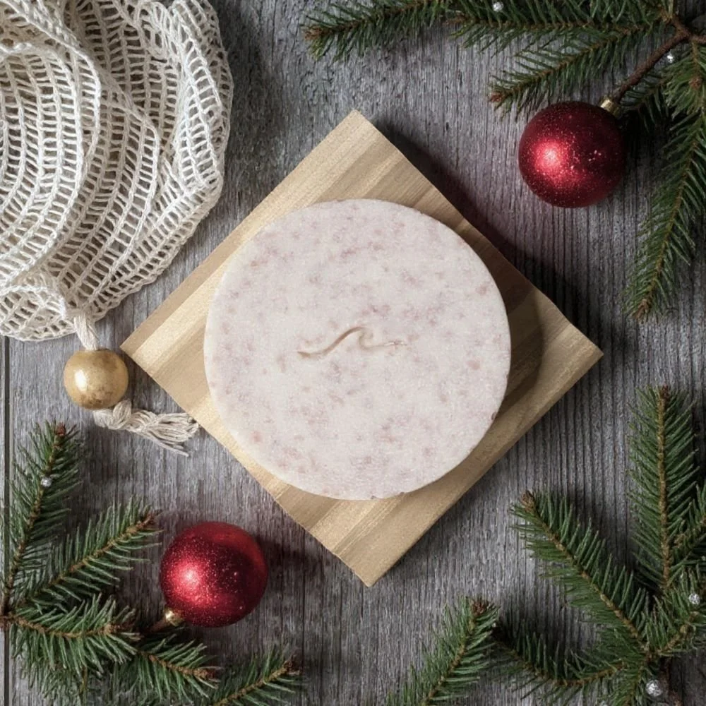 A round, pink and white soap bar on a wooden surface, surrounded by Christmas ornaments and pine branches.