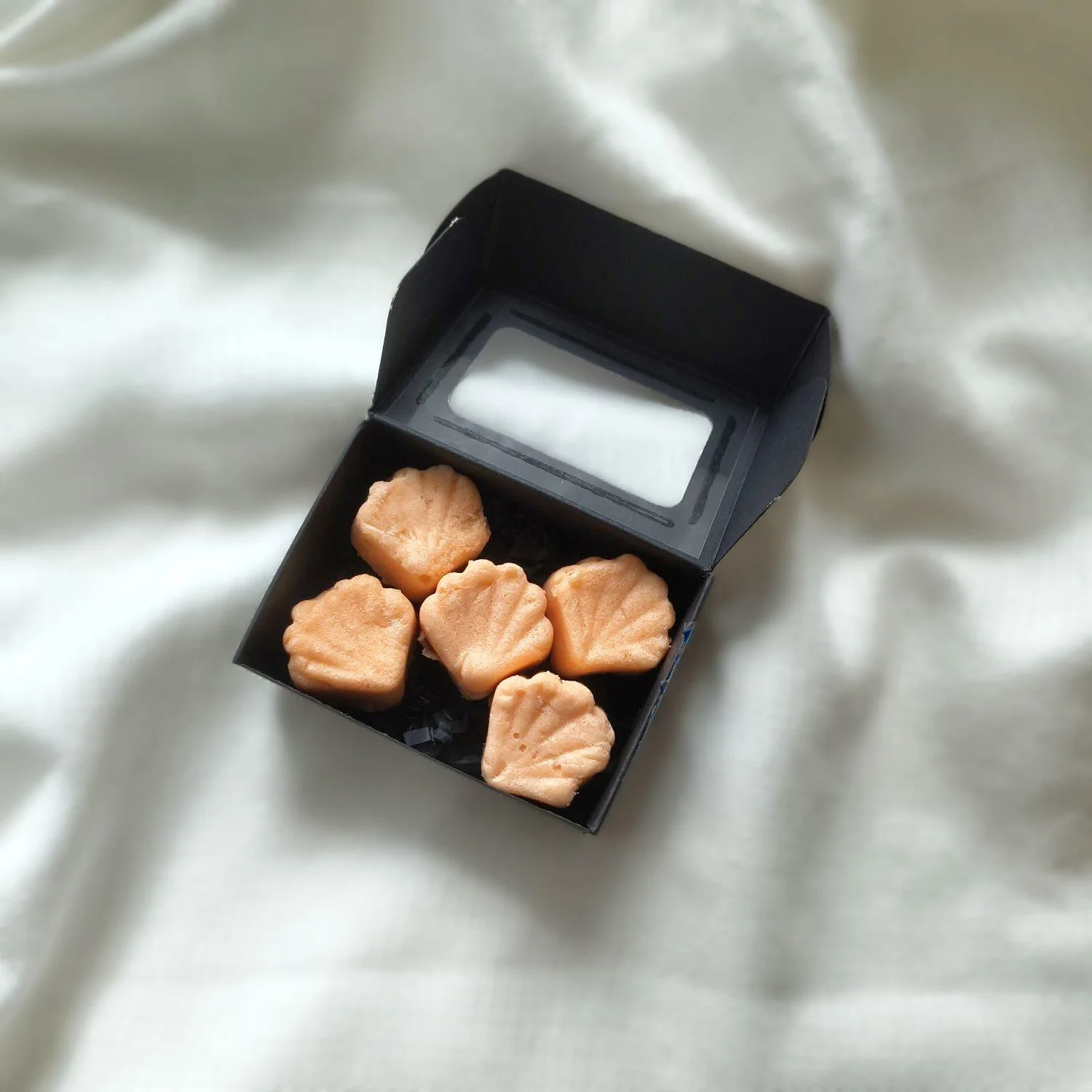 Set of five orange colored, shell shaped Pumpkin Santal Sugar Balm Shells in black box on textured cream background.