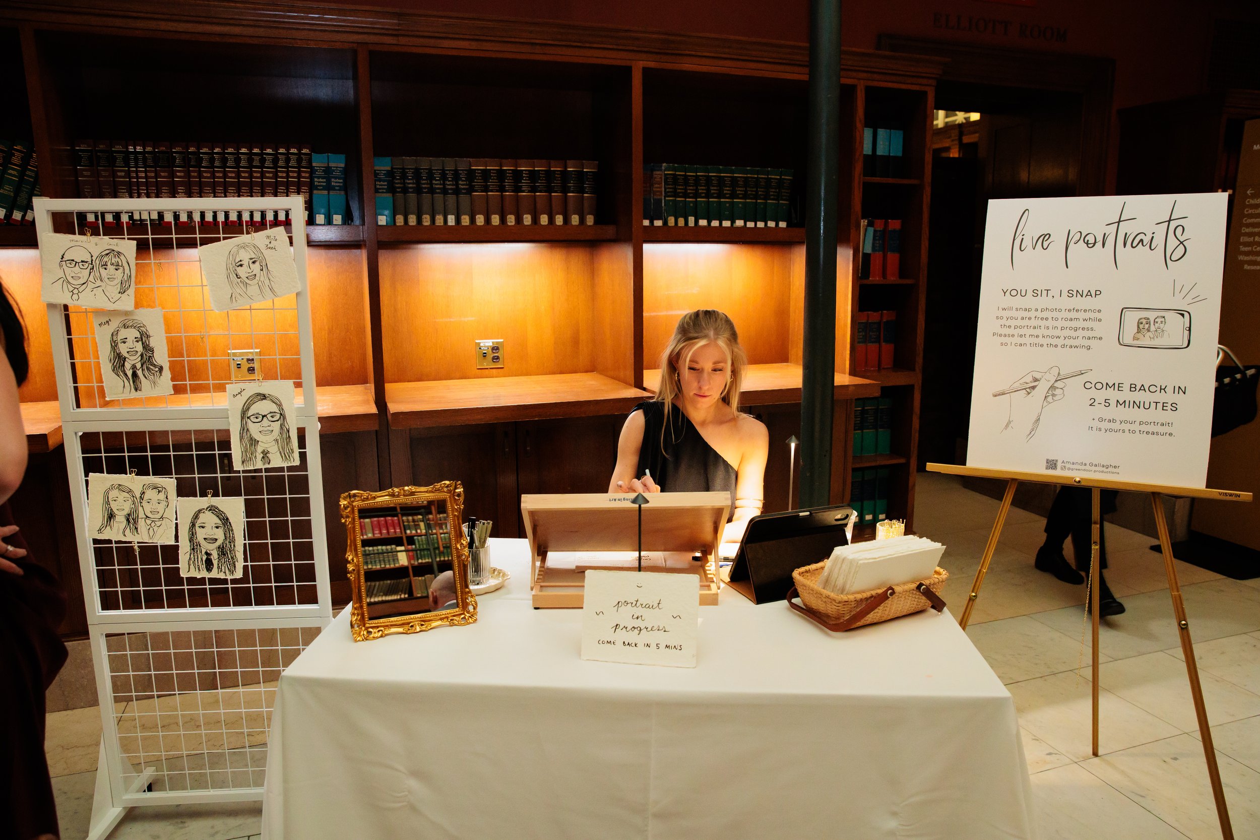 Amanda at a portrait drawing station with a sign and display of caricature-style portraits at Boston Public Library