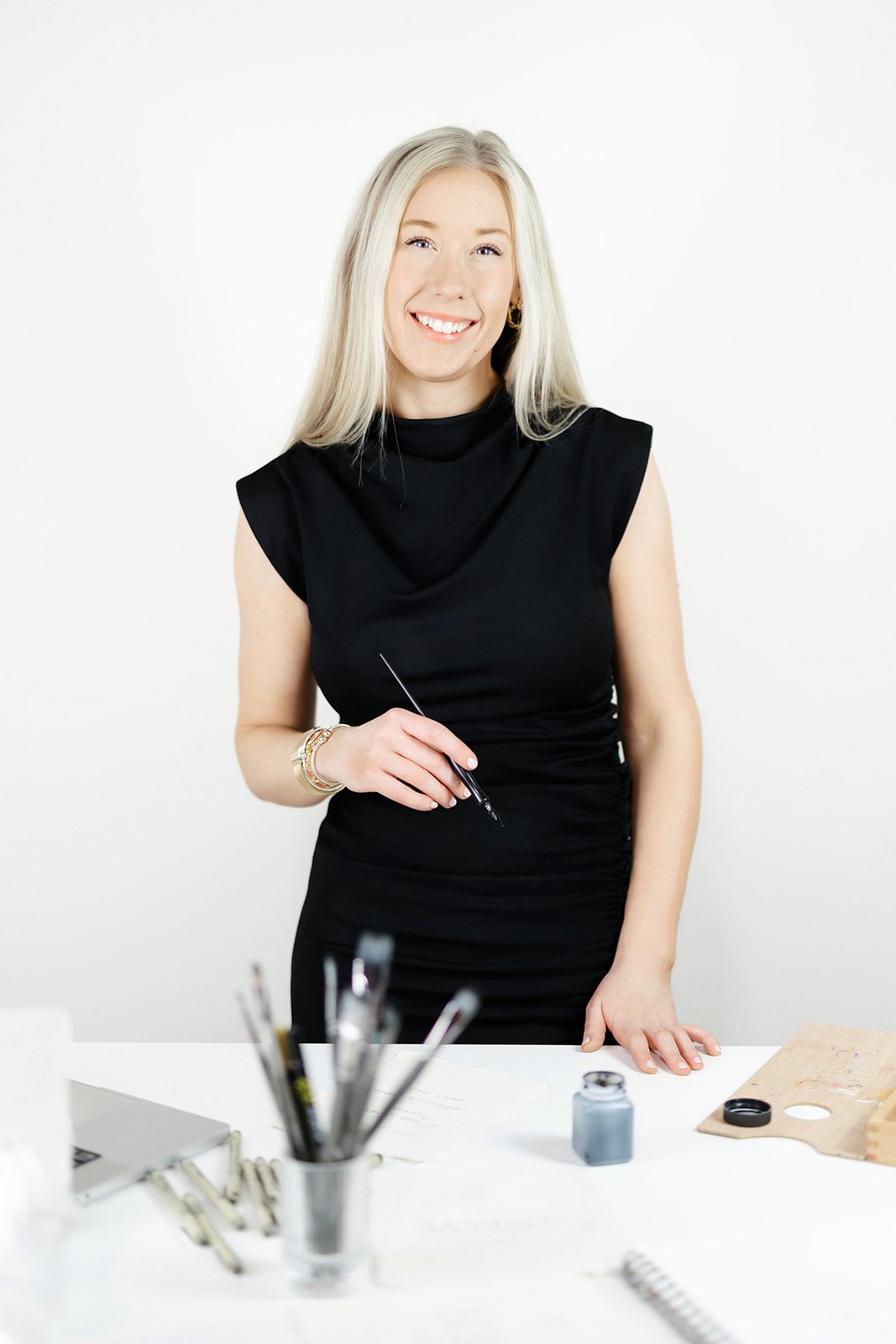 Amanda wearing a black dress, standing in front of a desk with art supplies, smiling and holding a brush.