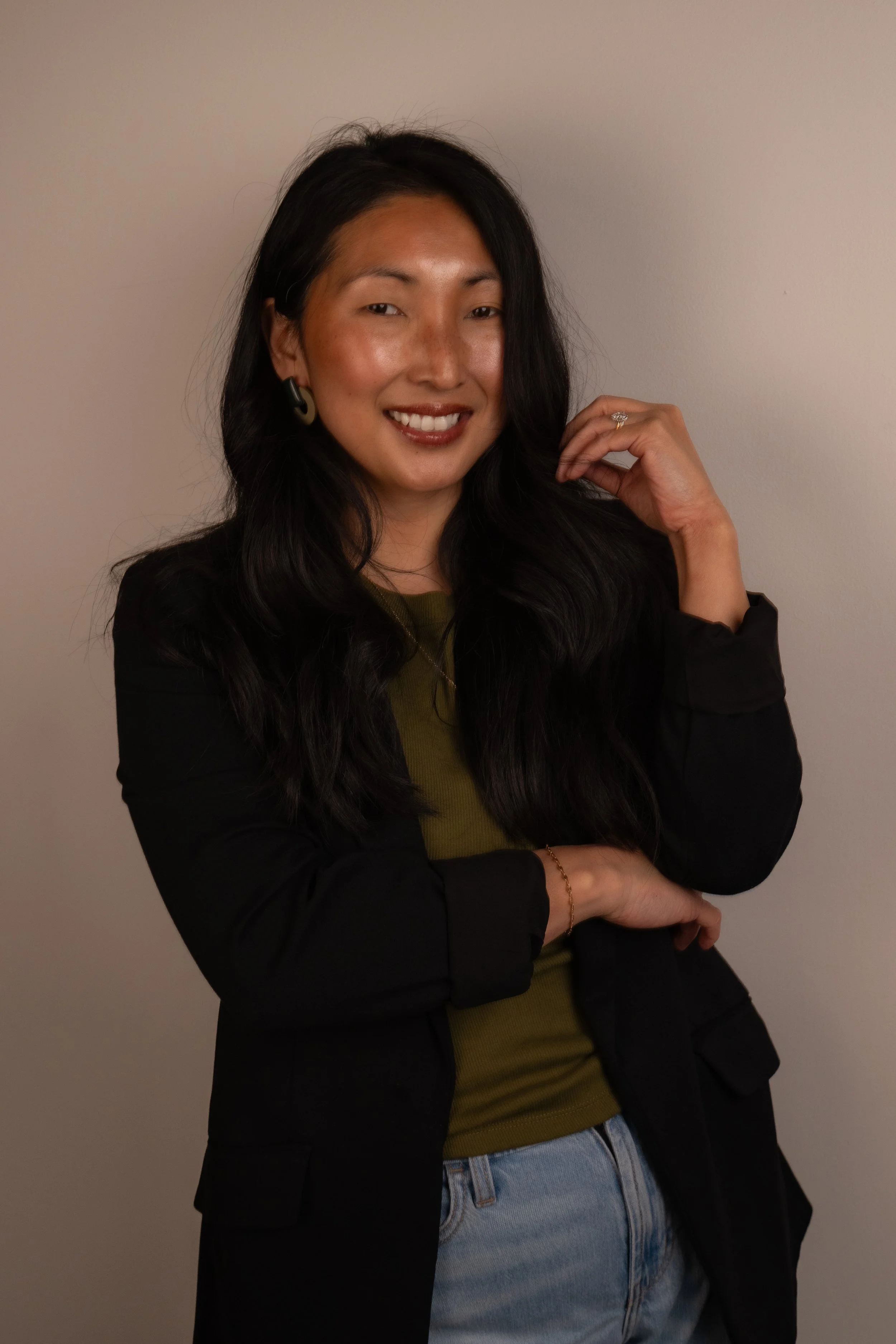 Person with black hair wearing a white blazer over a green top, posing against a plain background.