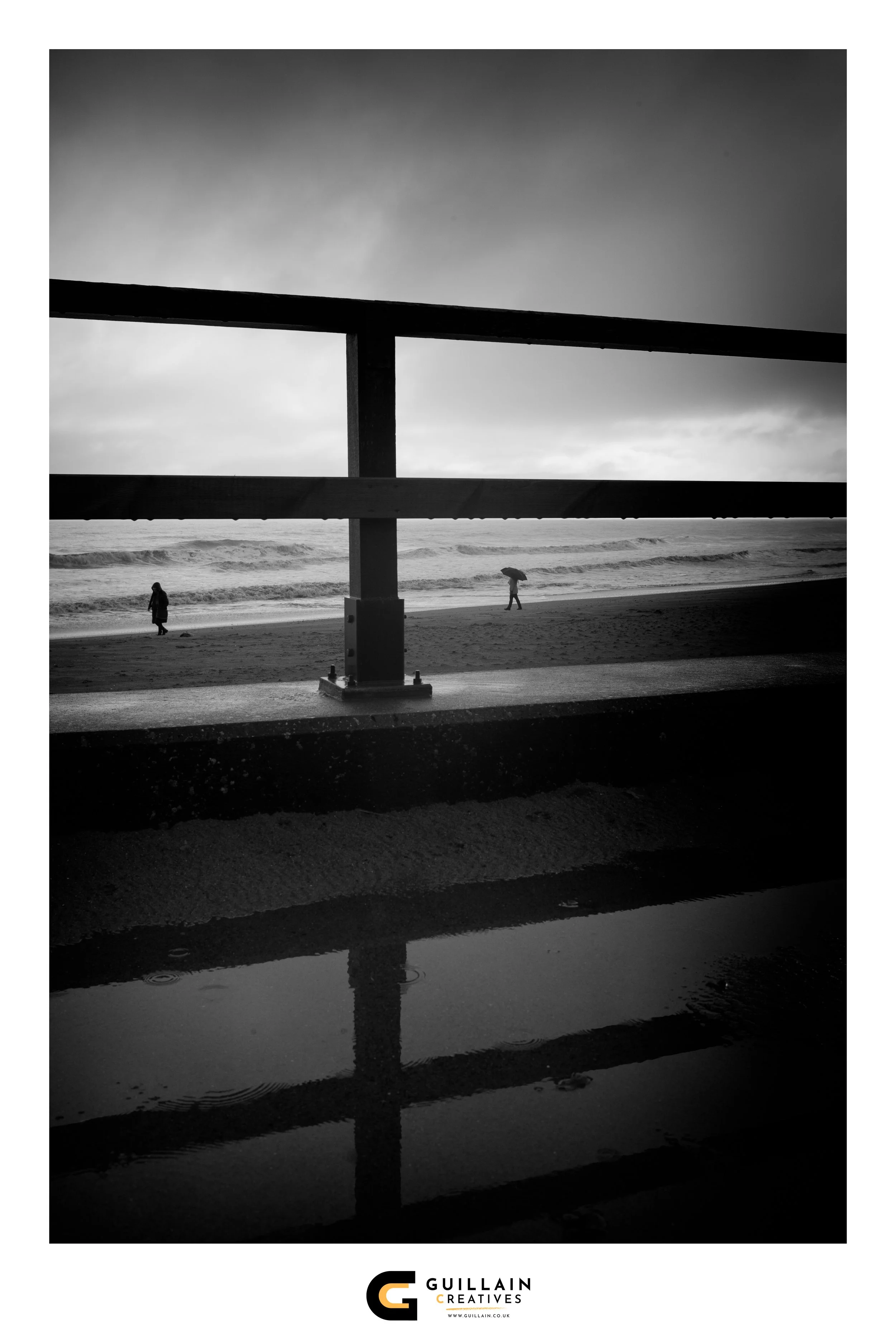 Black and white photo of a beach scene viewed through a railing, with three people walking near the water, one with an umbrella and two without, overcast sky, waves in the background, and a small pool of water in the foreground reflecting the railing