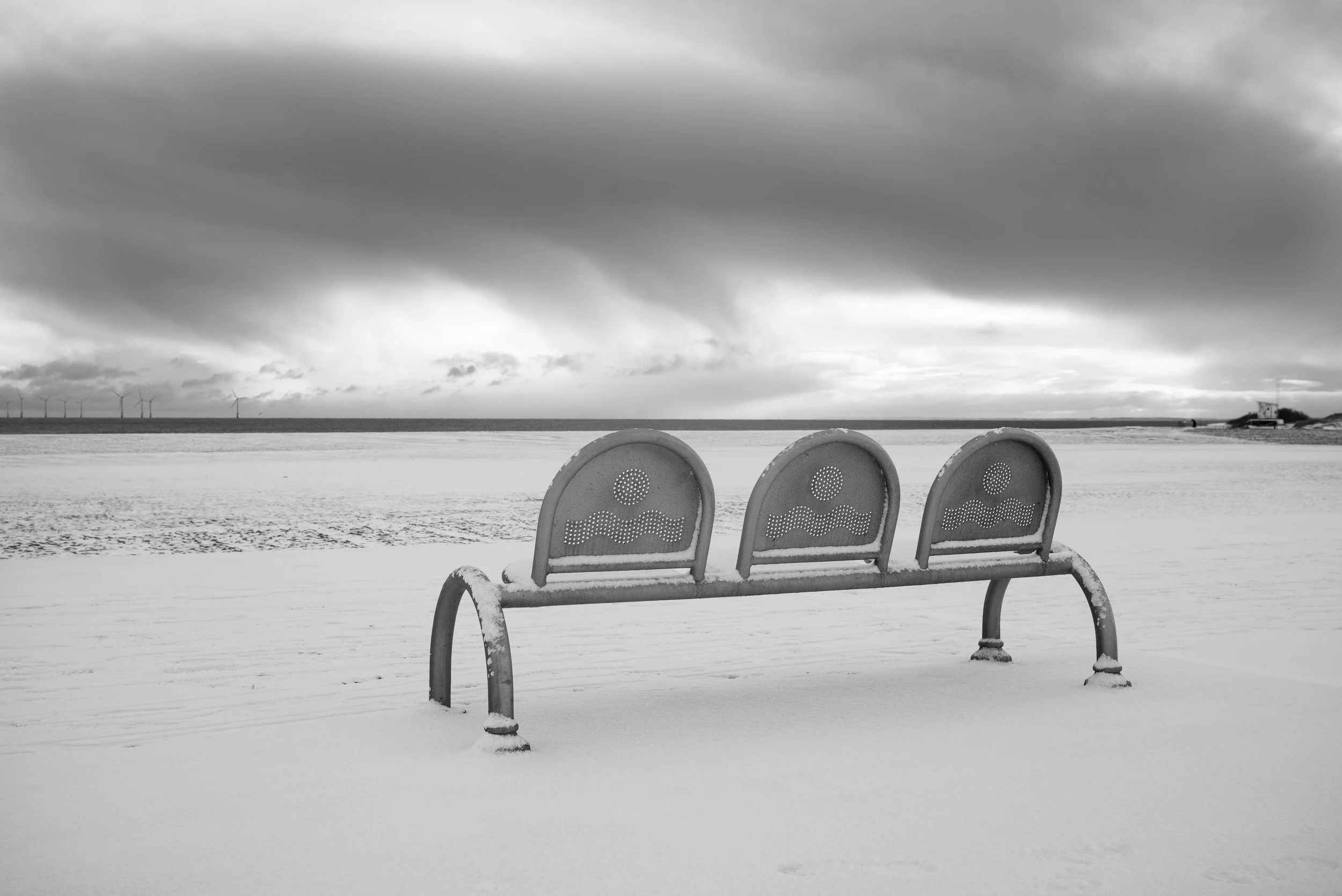 Empty snow-covered bench facing the ocean on a cloudy day.