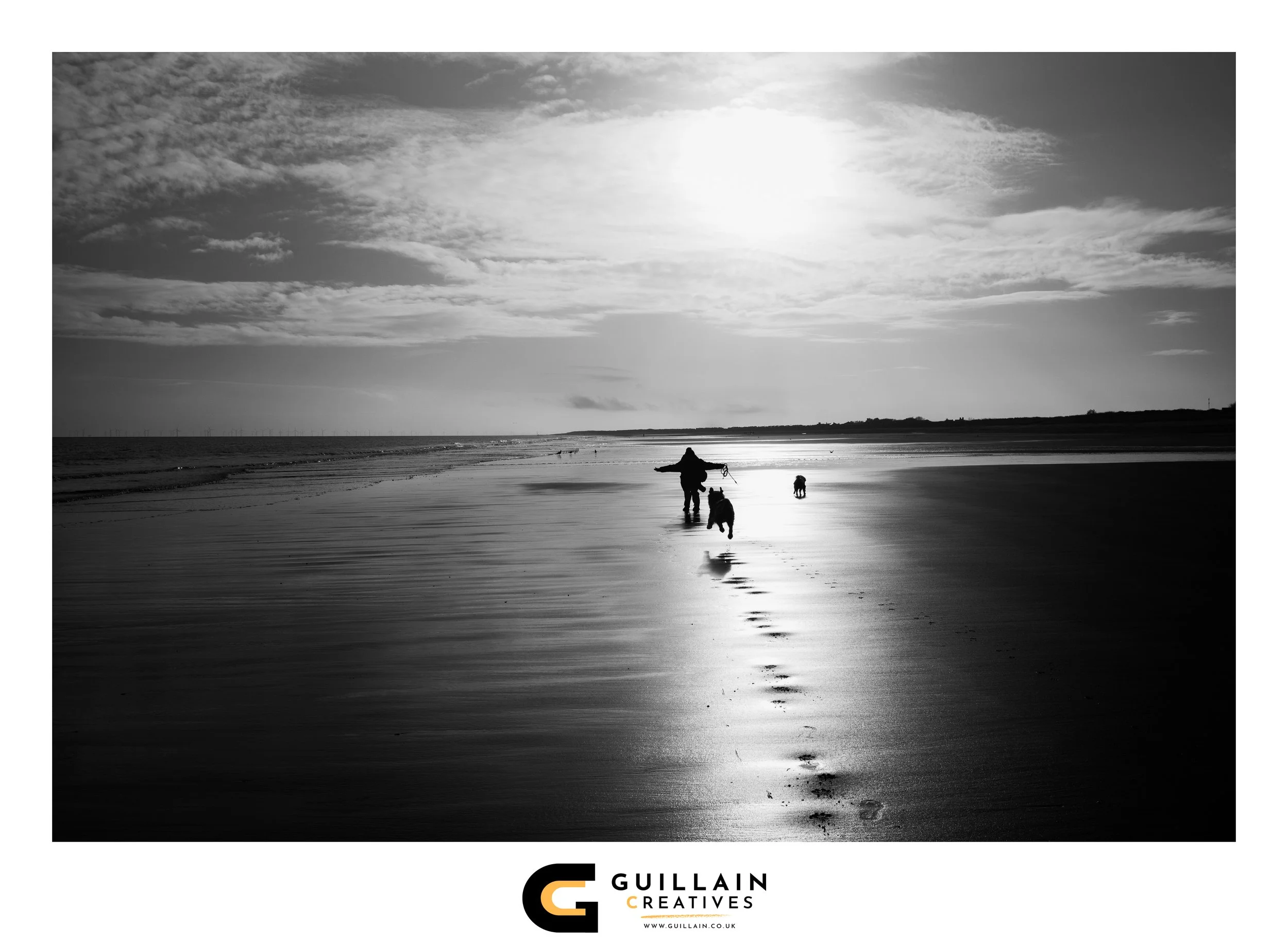 A person walking on the beach with two dogs during sunset, footprints leading from the foreground to the person, with a cloudy sky and calm ocean in the background.